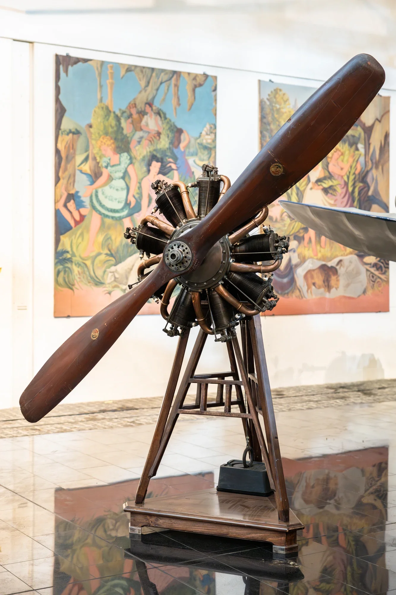 Une maquette d'avion en bois avec un moteur d'avion en cuivre et un grand rotor en bois, exposée sur un socle en bois dans un musée, avec des peintures colorées en arrière-plan.