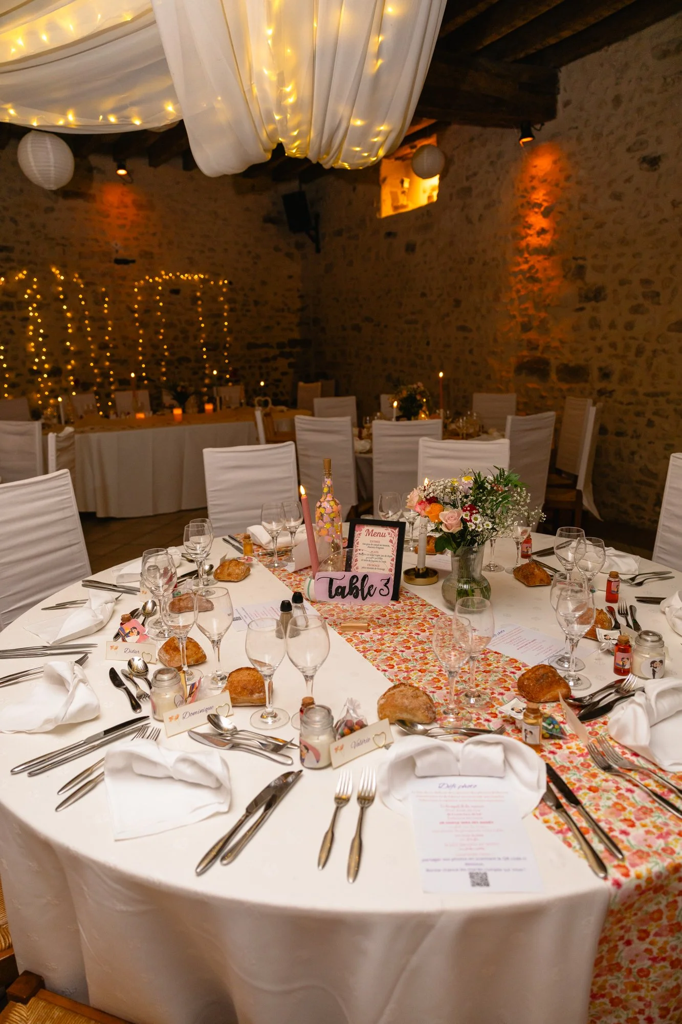 Une table décorée pour un mariage ou une fête, avec des assiettes, des verres, des couverts, des petits pains, une nappe de fleurs colorées, des bougies, un menu, des noms et des petits cadeaux. La pièce a des murs en pierre avec des lumières chaleur