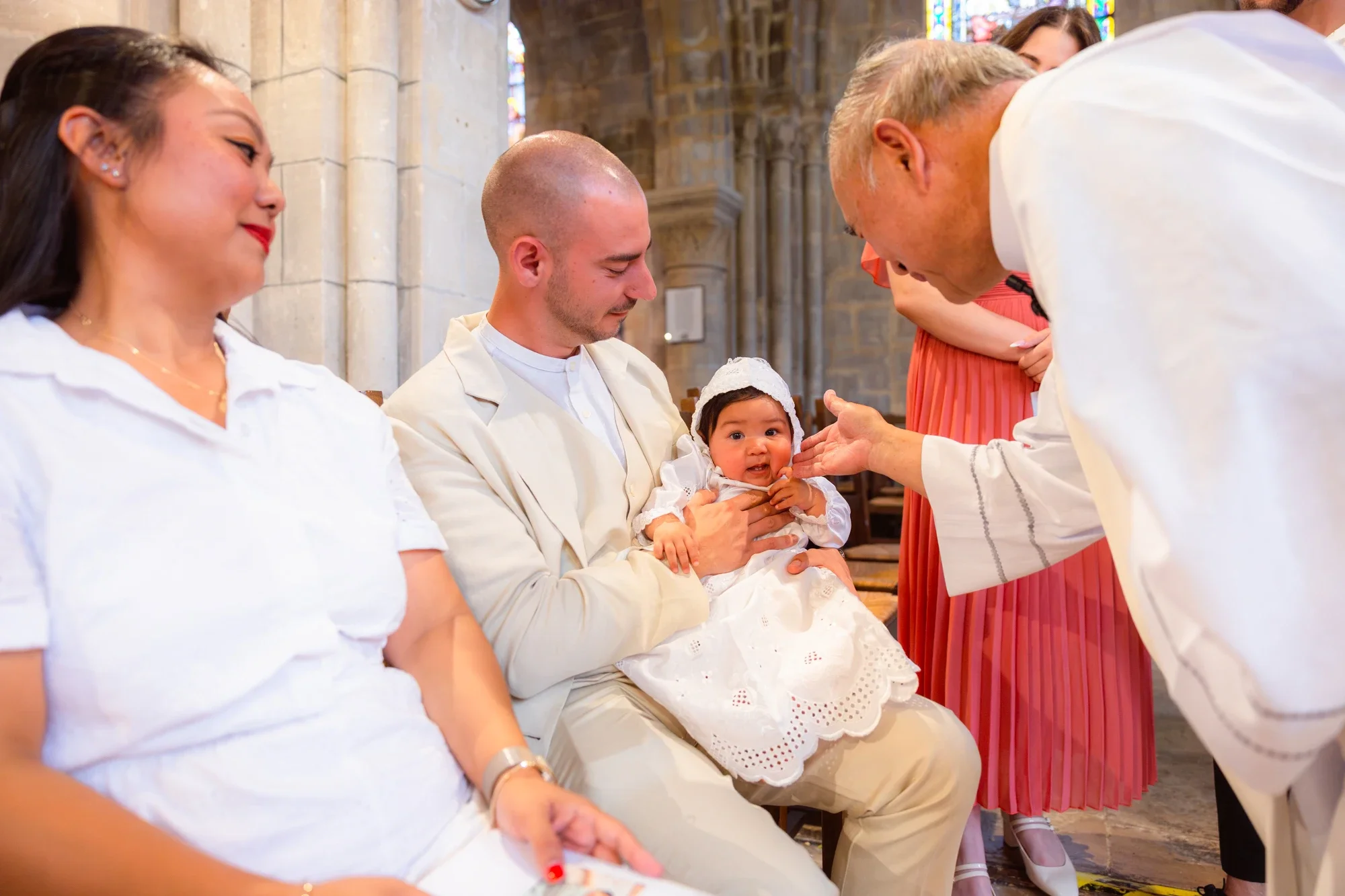 Un bébé est baptisé dans une église tandis qu'un groupe de personnes assises attendent dans la nef, avec des vitraux colorés en arrière-plan.