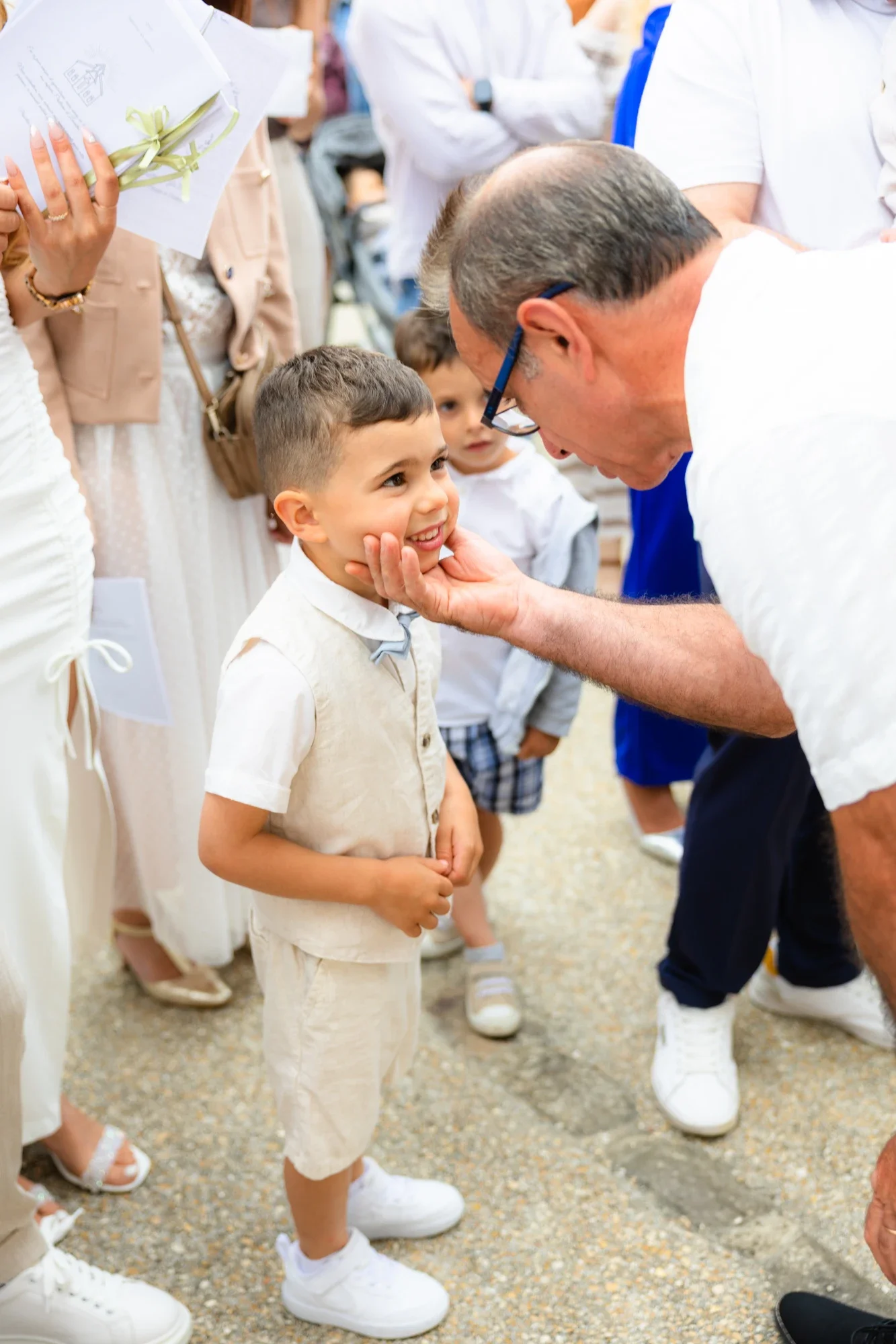 Un garçon souriant, habillé en beige avec une veste et un pantalon en lin, regarde un homme âgé lui caresser la joue lors d'un événement, entouré d'autres enfants et adultes, dans une ambiance festive et élégante.