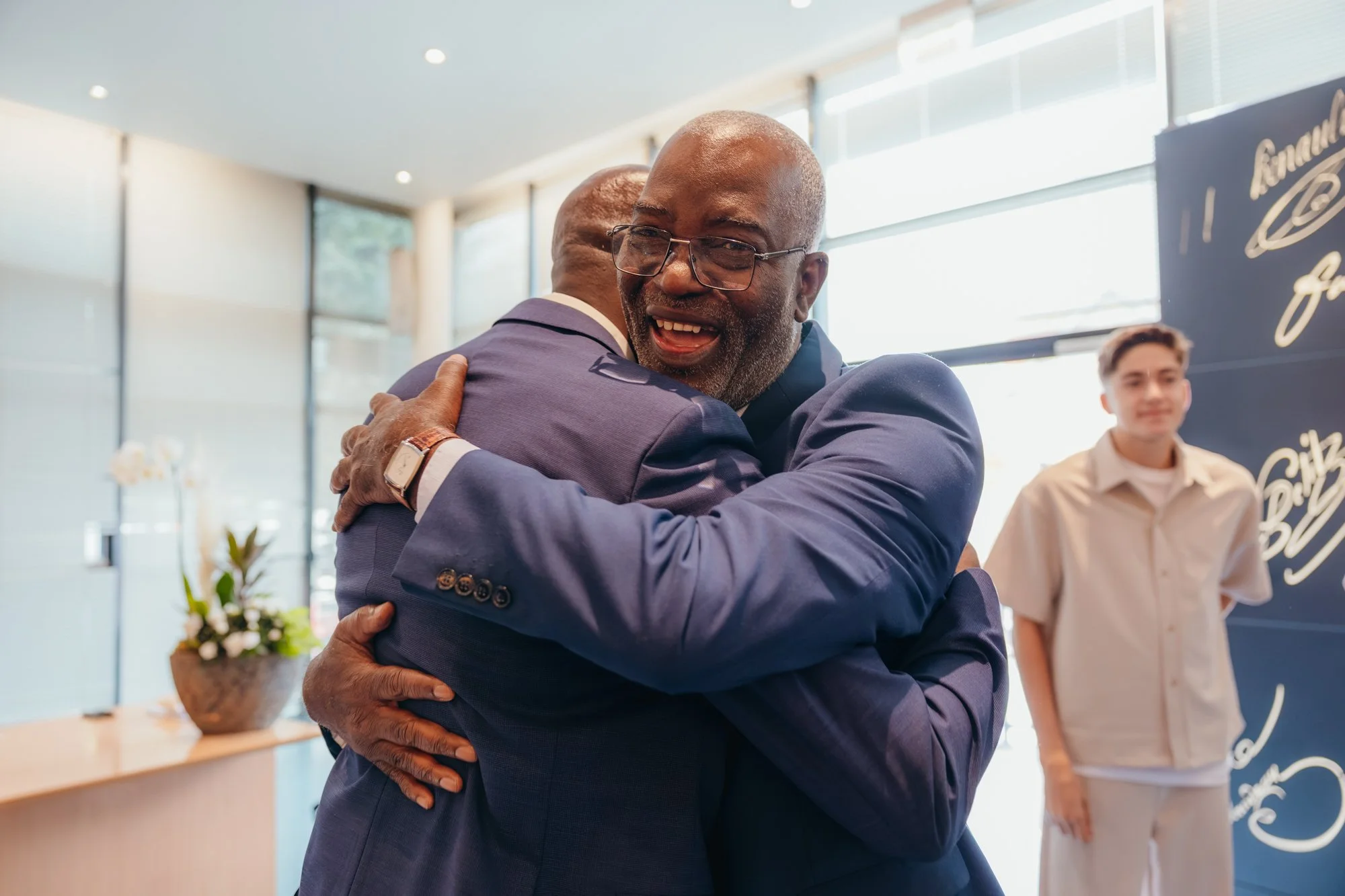 Deux hommes d'affaires souriants se serrent la main lors d'une accolade chaleureuse dans un lieu lumineux, peut-être à une conférence ou un événement professionnel. En arrière-plan, un jeune homme se tient près d'un panneau avec des dessins et textes