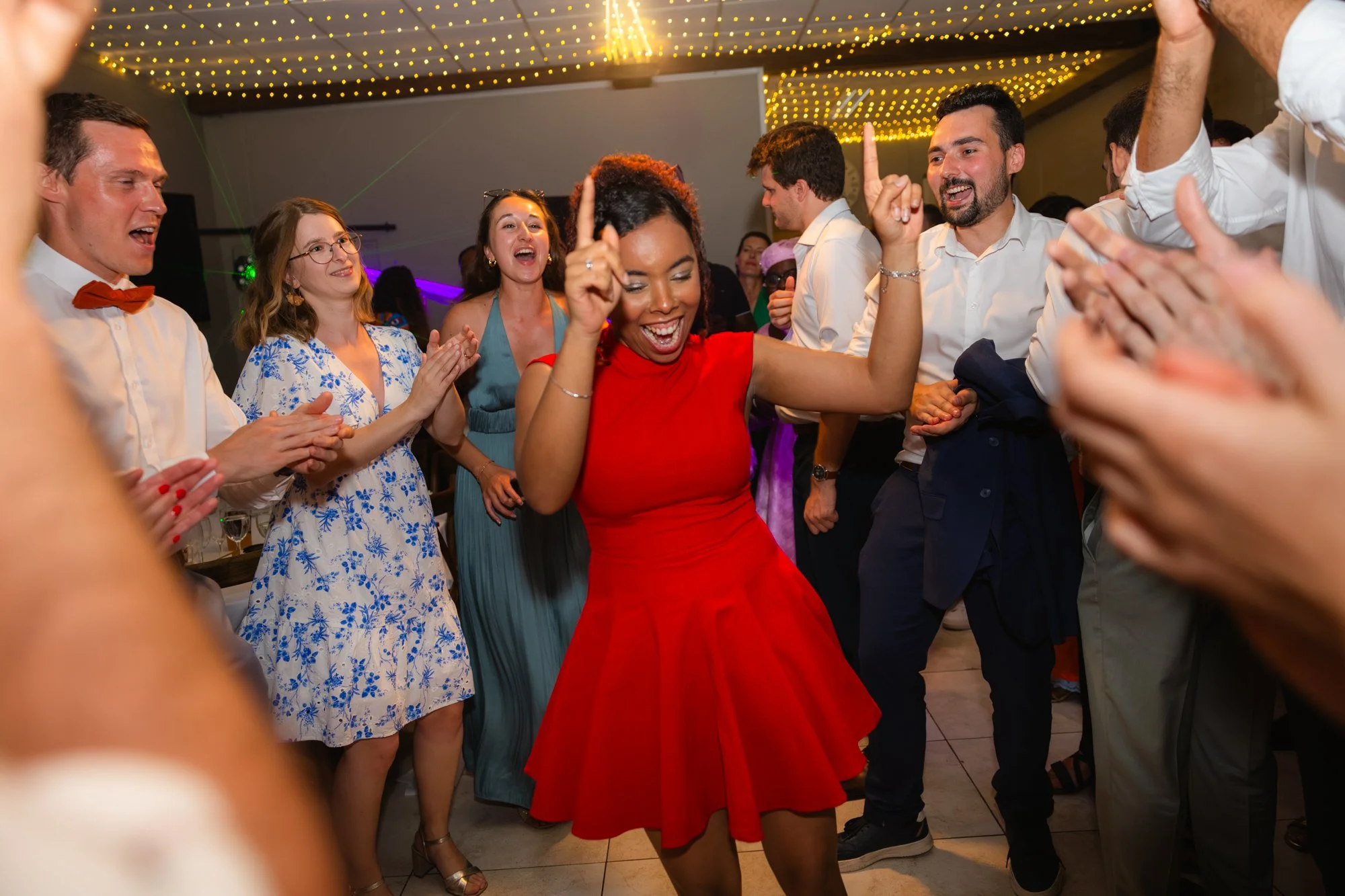 un groupe de personnes dans une fête dansent et s'amusent, notamment une femme en robe rouge qui se déhanche, entourée d'autres personnes qui applaudissent et rient, sous un plafond décoré de guirlandes lumineuses.