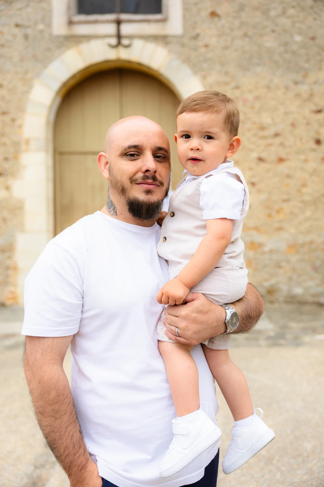 Un homme musclé avec une barbe et un tatouage sur le cou, portant un t-shirt blanc, tient un jeune garçon dans ses bras. Le garçon porte une chemise blanche, un short beige et des chaussures blanches. Ils sont devant un bâtiment en pierre avec une gr