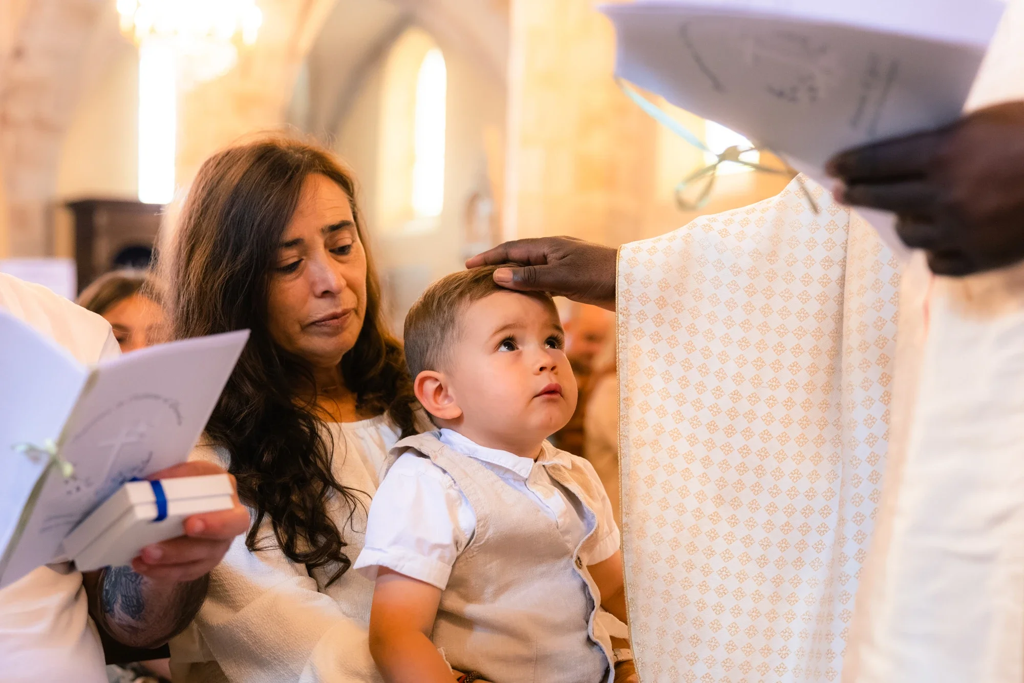Enfant et adulte religieux pendant une cérémonie religieuse, l'adulte pose sa main sur la tête de l'enfant qui regarde attentivement. La scène se déroule dans un lieu de culte avec une lumière chaleureuse et un décor en pierre de fond.