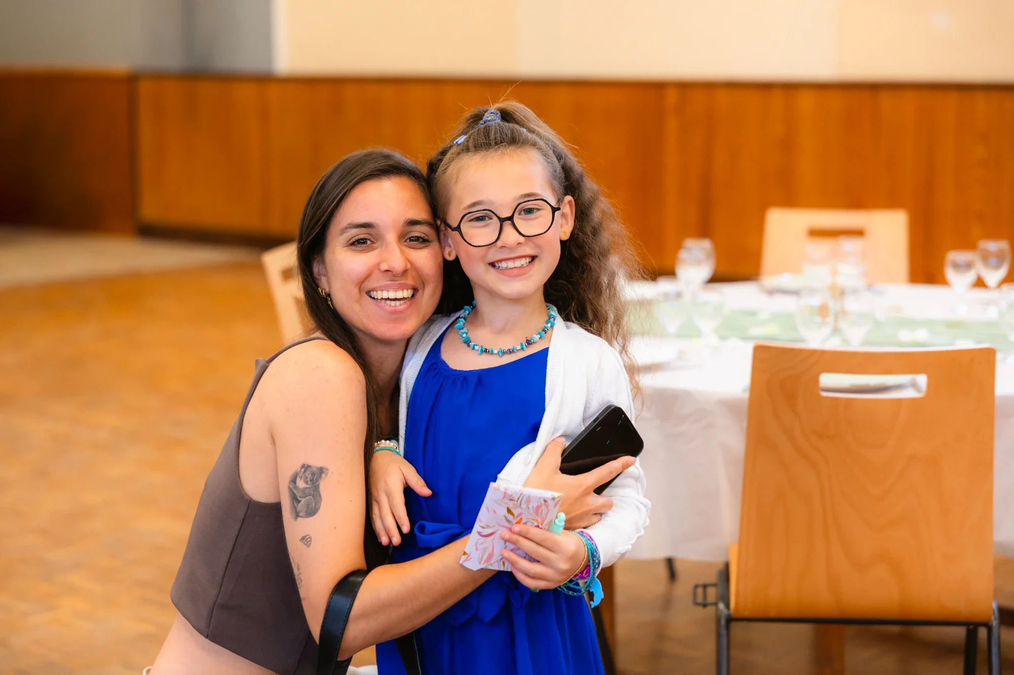 Une femme et une jeune fille souriantes posent ensemble dans une salle à manger. La femme a des cheveux bruns, portant un haut gris, et la jeune fille a des cheveux bouclés, portant des lunettes, une robe bleue et des bijoux colorés.