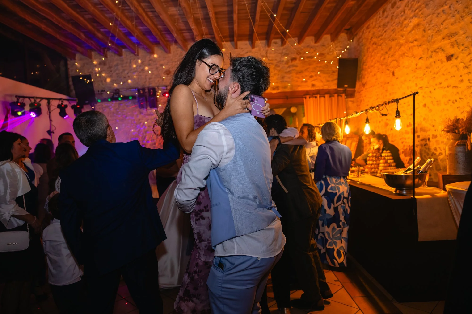 Un couple danse et s'embrasse lors d'une fête dans une salle avec murs en pierre et éclairage tamisé, décorée avec des guirlandes lumineuses.