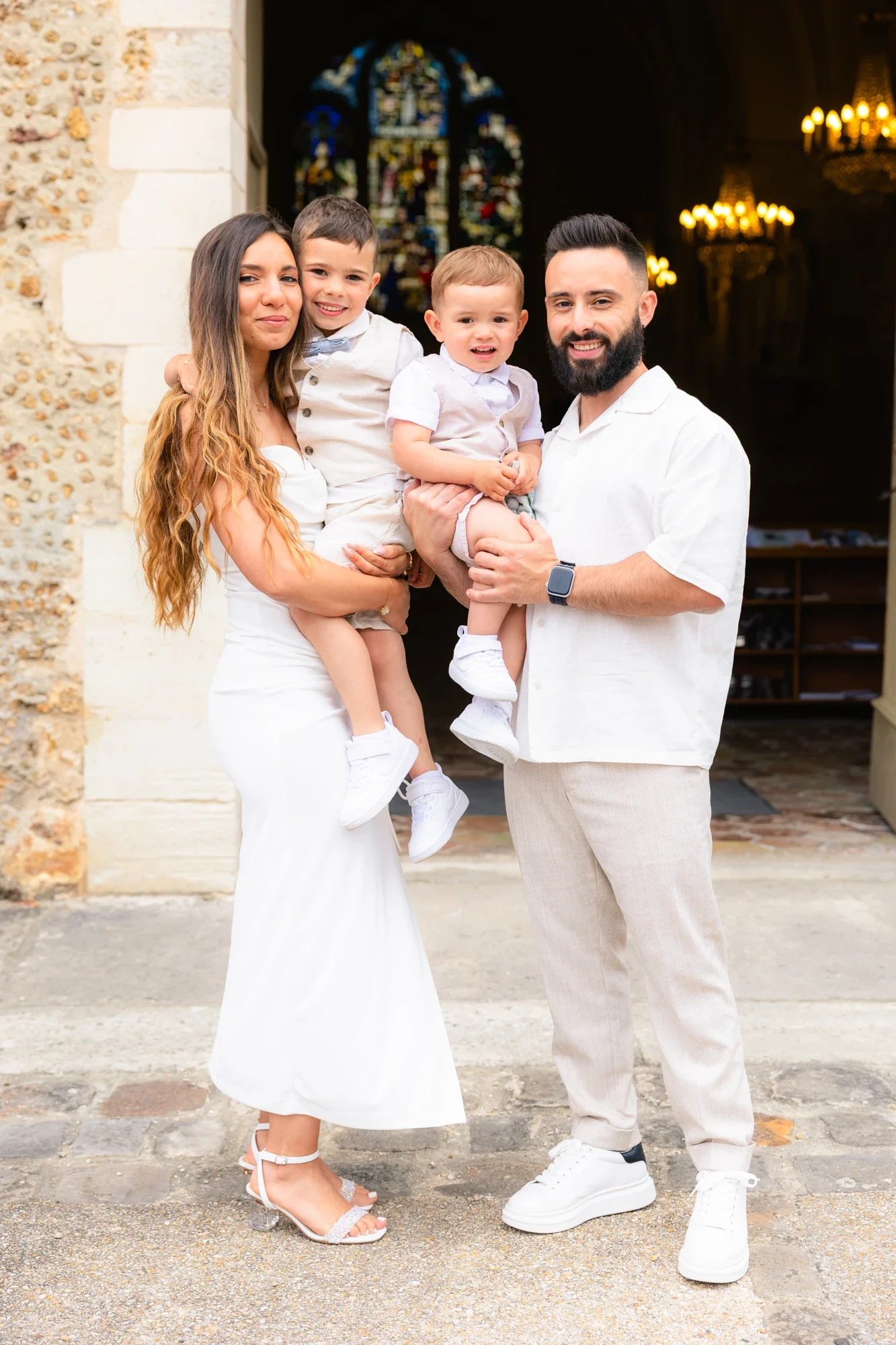 Une famille pose devant une église, la mère porte une robe blanche et des chaussures à talons, le père porte une chemise blanche et un smartwatch, les deux enfants portent des vêtements blancs et sont tenus dans les bras de leurs parents.