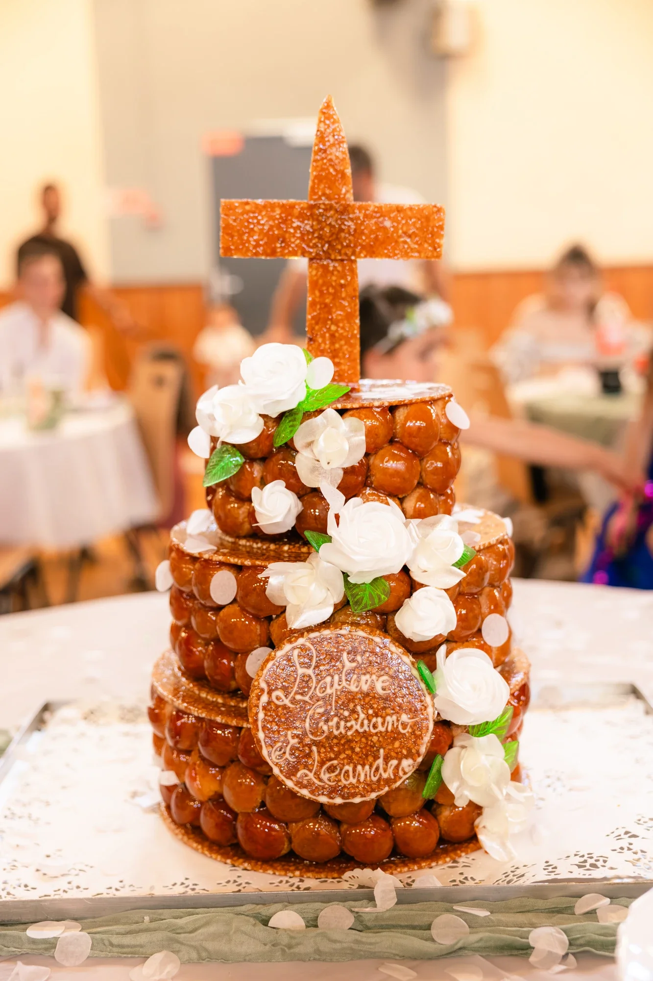 Gâteau de mariage en forme de croix avec décor de fleurs blanches et un disque en biscuit portant l'inscription "Love, Catholic, Alexandre".