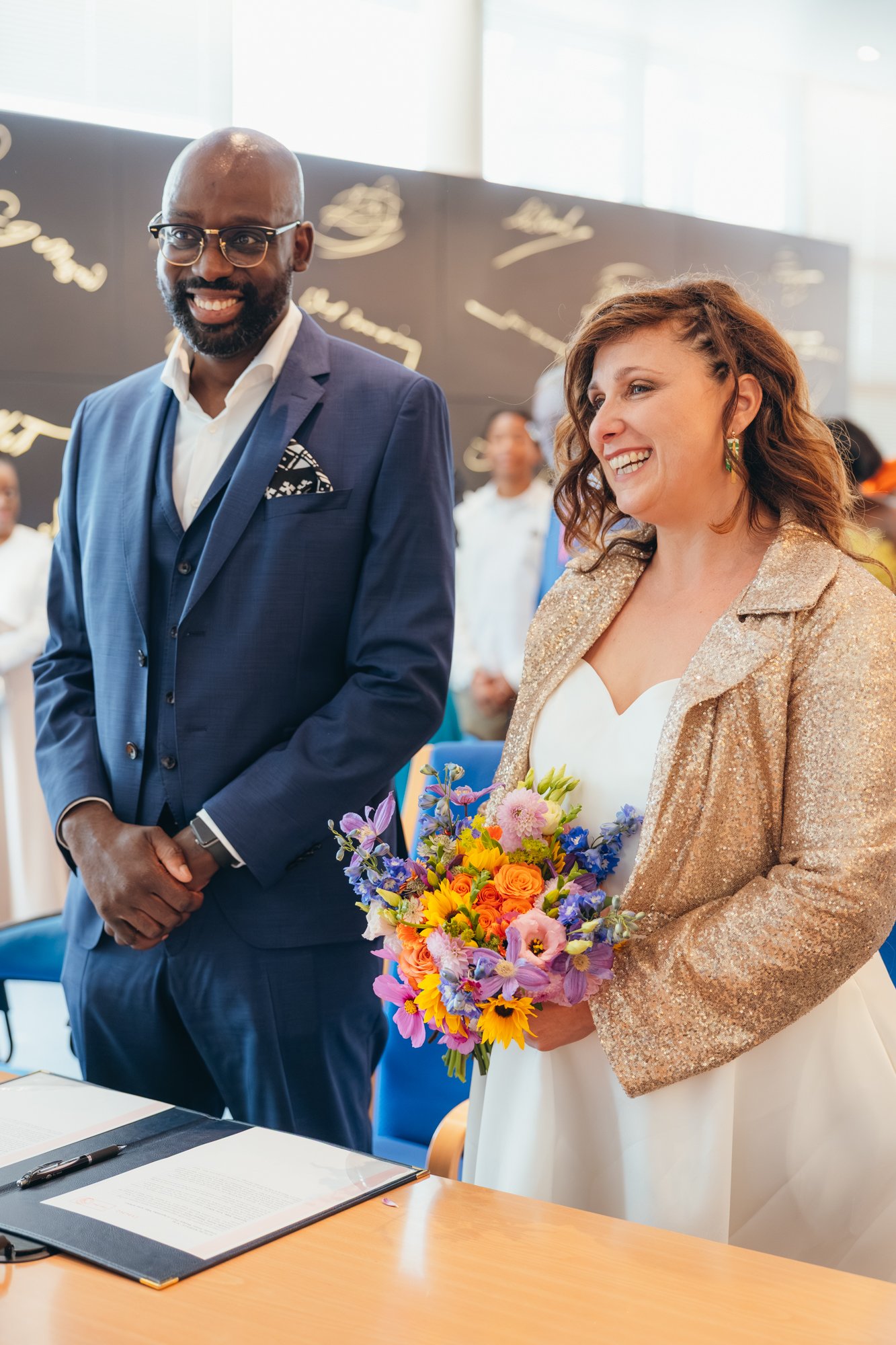 Un homme d'affaires en costume bleu et une femme habillée en blanc avec une veste or, tenant un bouquet de fleurs, lors d'une cérémonie en intérieur.
