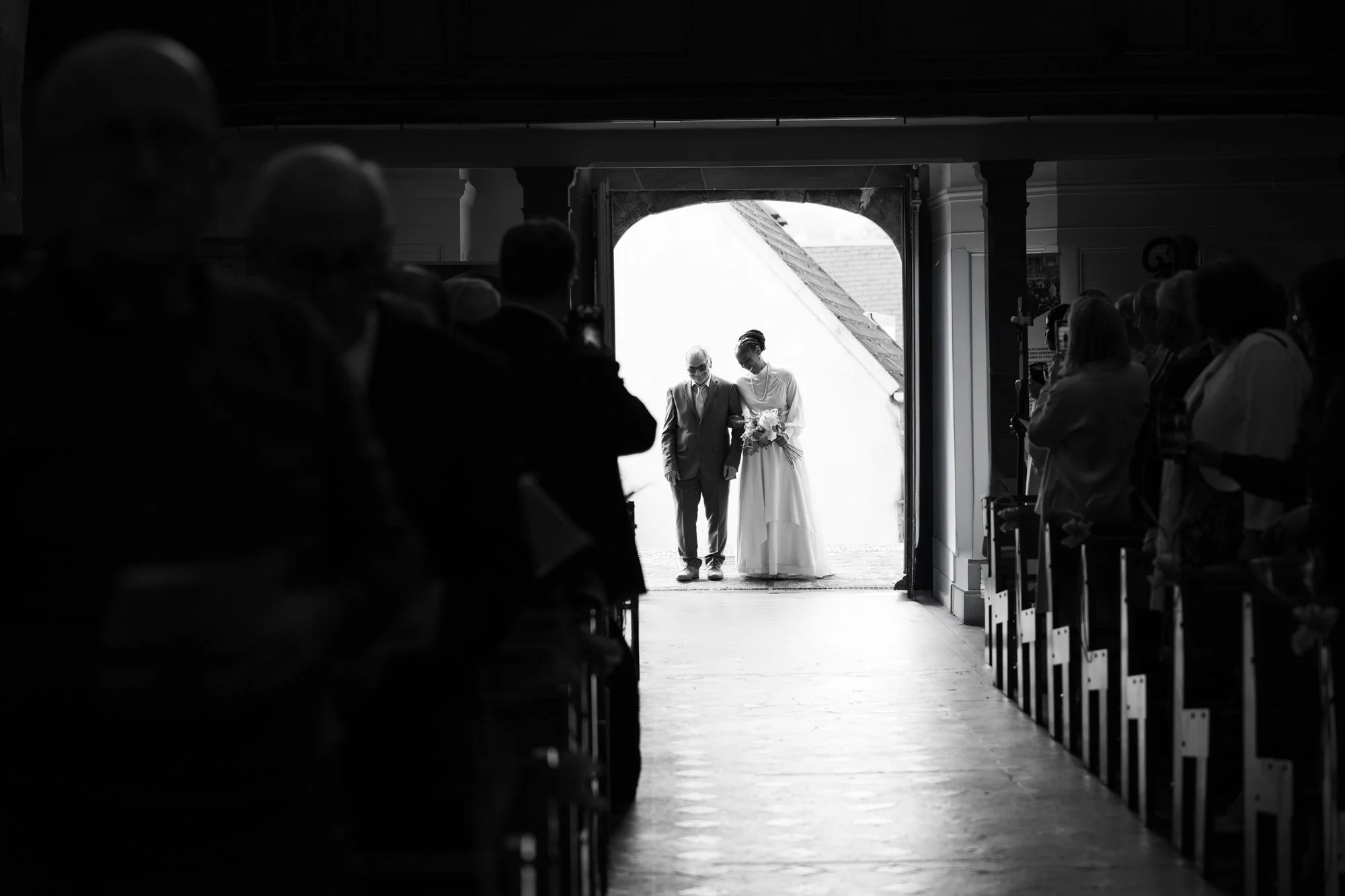 Un couple de mariés marche vers la sortie d'une église, entouré de nombreux invités. L'image est en noir et blanc, avec la luminosité en arrière-plan.