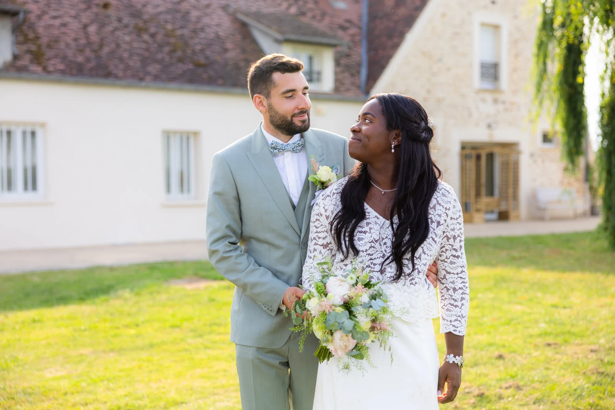Un couple de mariage, un homme en costume gris avec un noeud papillon et une femme en robe blanche tenant un bouquet, se regardent tendrement dans un jardin ensoleillé.