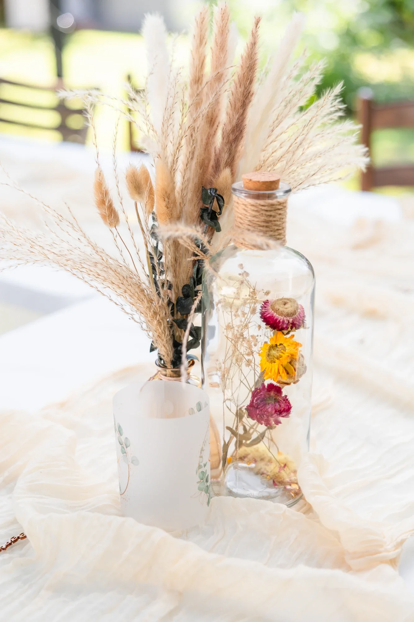 Une décoration de fleurs séchées dans des bocaux en verre sur une table couverte d'une nappe blanche.