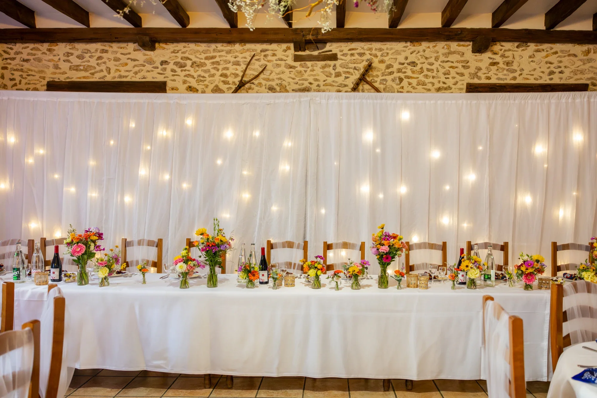 Table de fête décorée avec des bouquets de fleurs colorées, bouteilles de vin et verres, dans une salle avec une décoration rustique, des rideaux lumineux derrière, et un plafond en bois.
