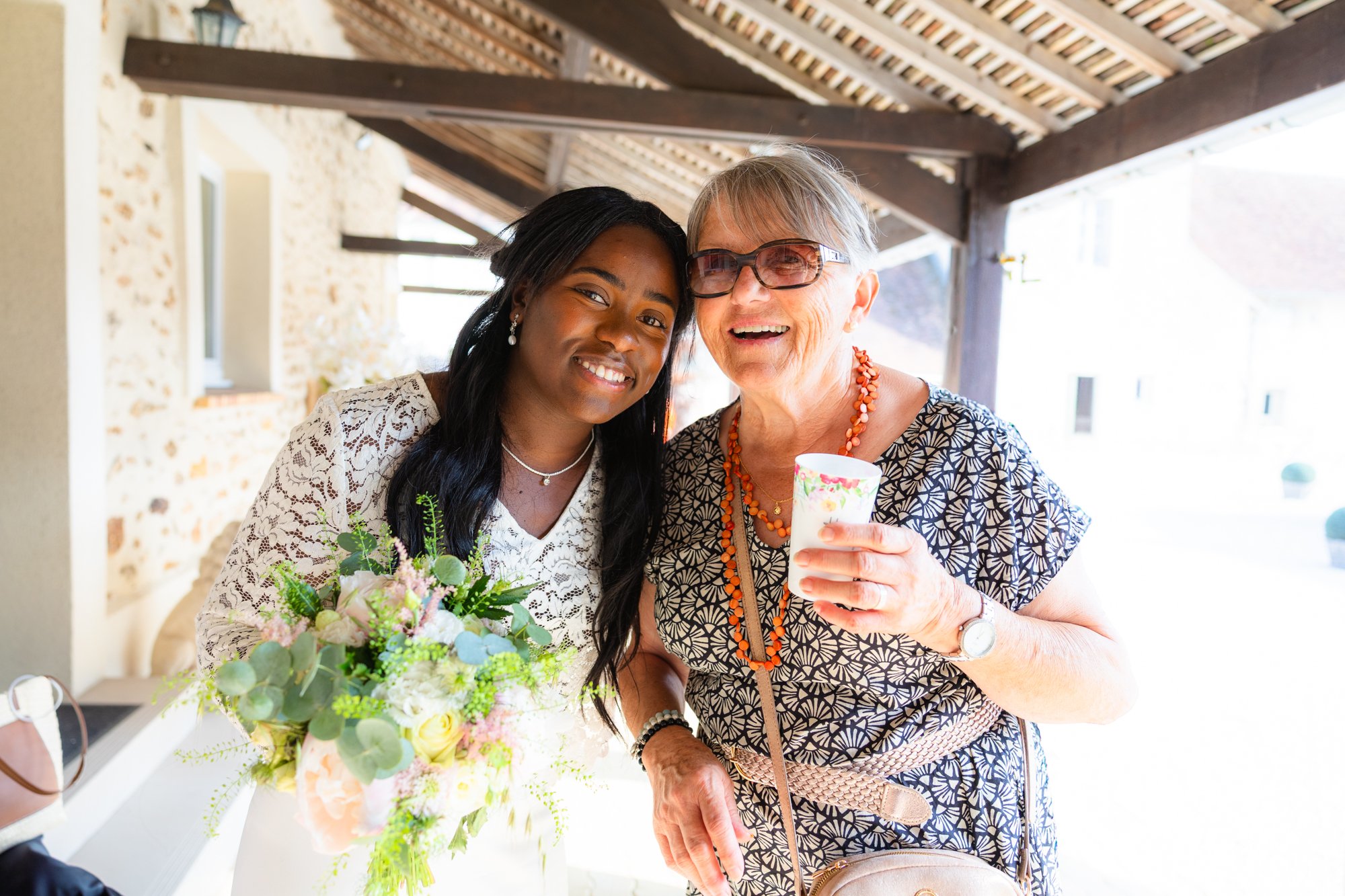 Deux femmes souriantes, une plus jeune et une plus âgée, posent ensemble dans un cadre en plein air sous un toit en bois, avec une table de fleurs devant elles, célébrant probablement un événement joyeux.