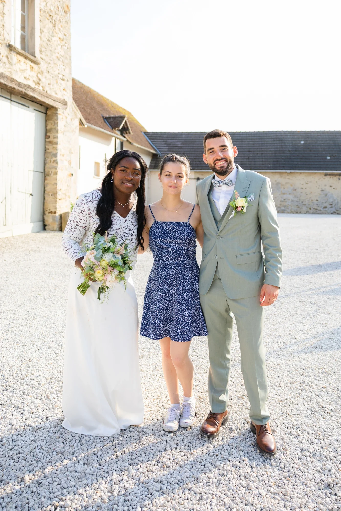 Trois personnes posent ensemble lors d'une célébration en plein air, avec une femme en robe blanche tenant un bouquet, une femme en robe bleue à pois et un homme en costume gris avec une fleur de boutonnière.
