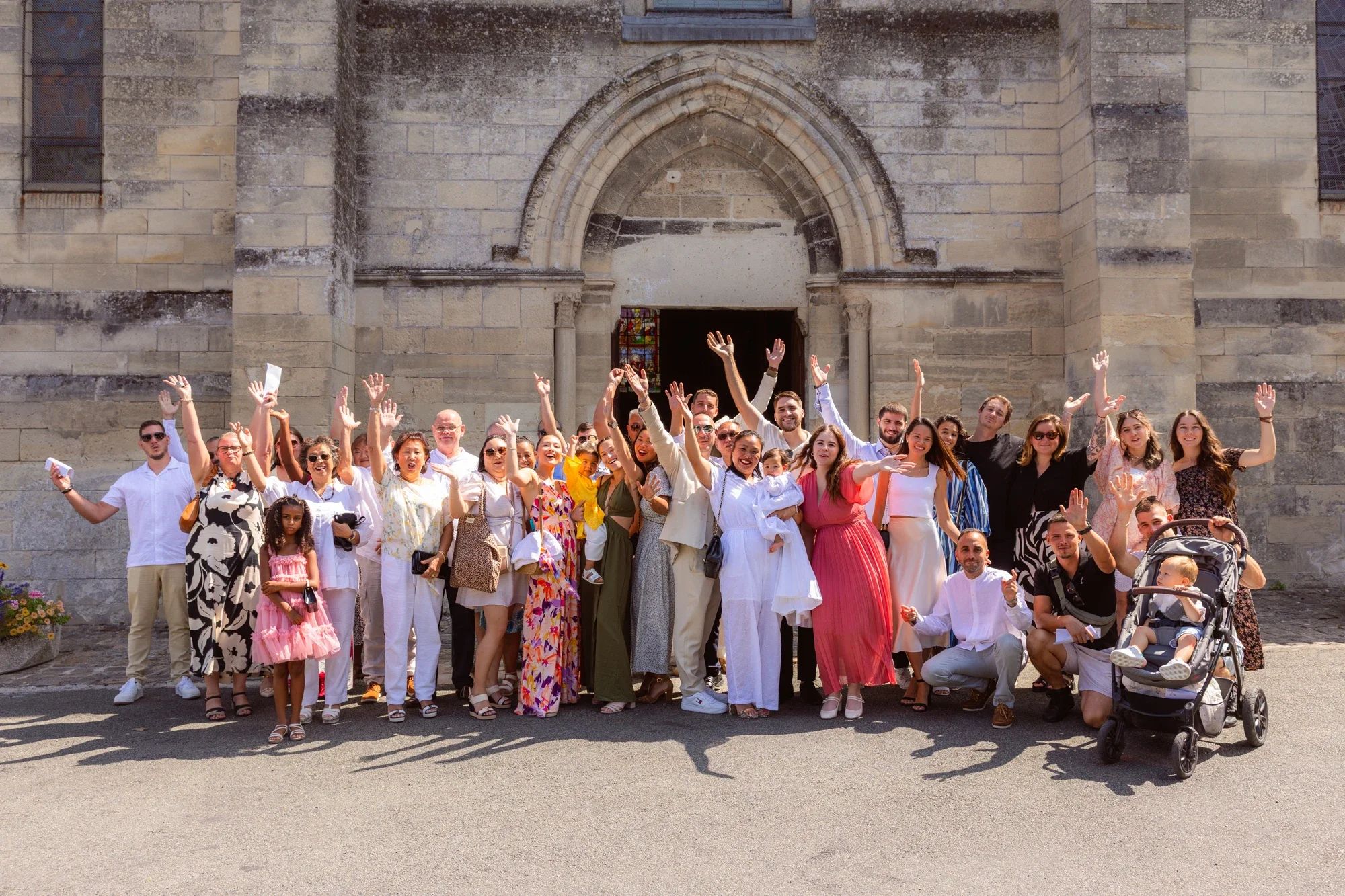 Groupe de personnes souriantes et saluant devant une vieille église en pierre, certains avec des enfants, un bébé, et une poussette, ambiance joyeuse et ensoleillée.