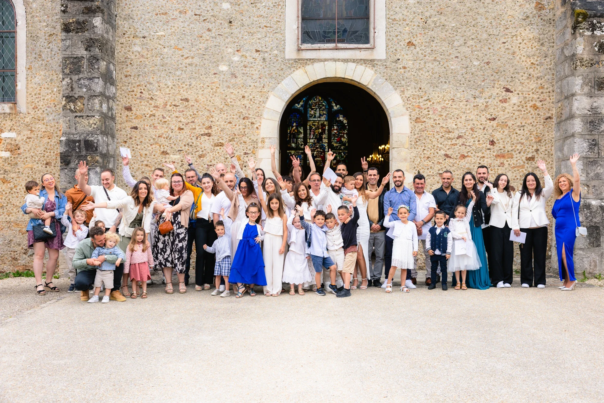 Groupe de personnes posant devant une vieille église en pierre avec un vitrail coloré et une fenêtre en métal. Certaines personnes portent des vêtements formels ou traditionnels, et toutes semblent sourire et saluer.