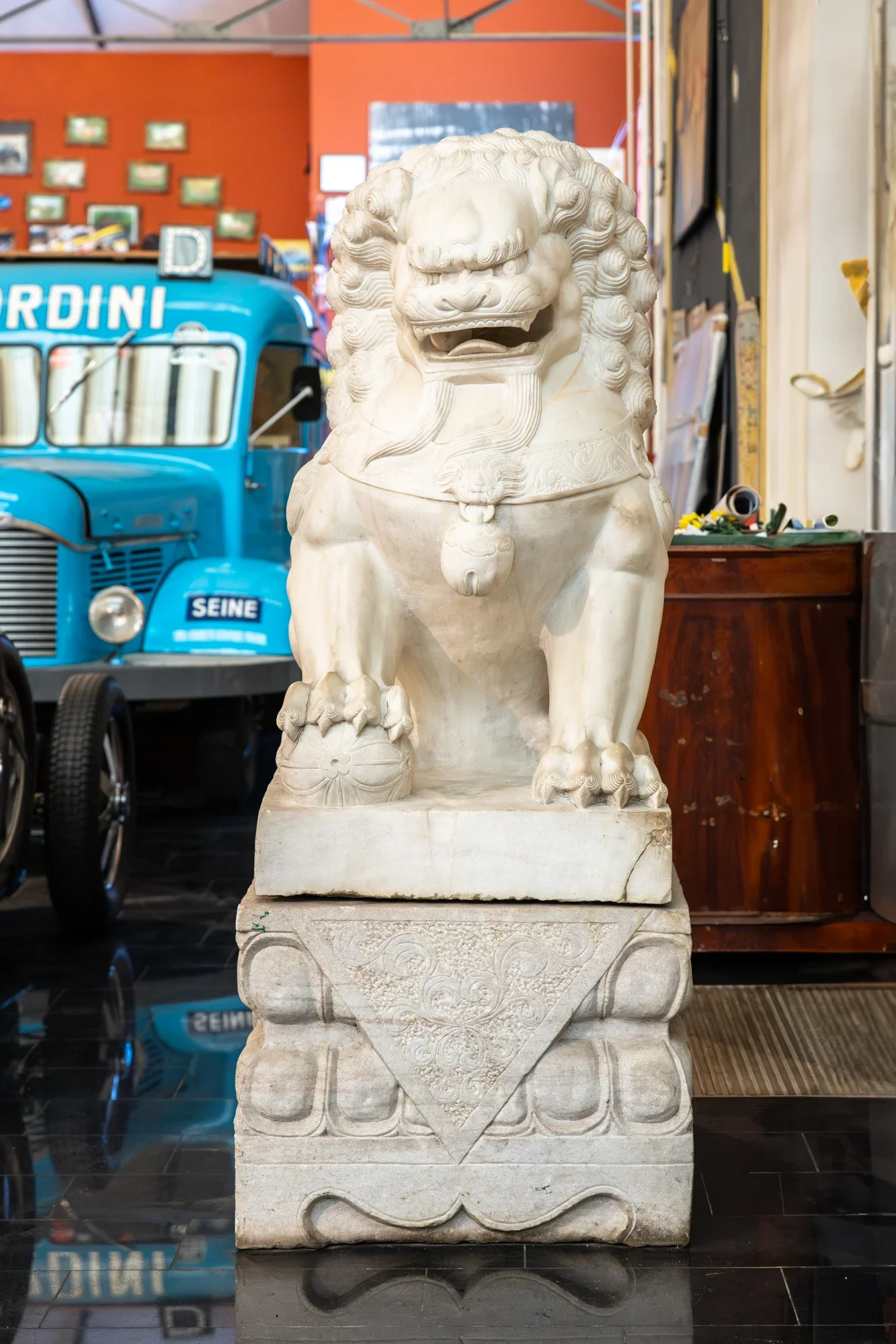 Statue de lion en pierre blanc exhibée dans une galerie ou un musée avec fond orange et décorations murales.