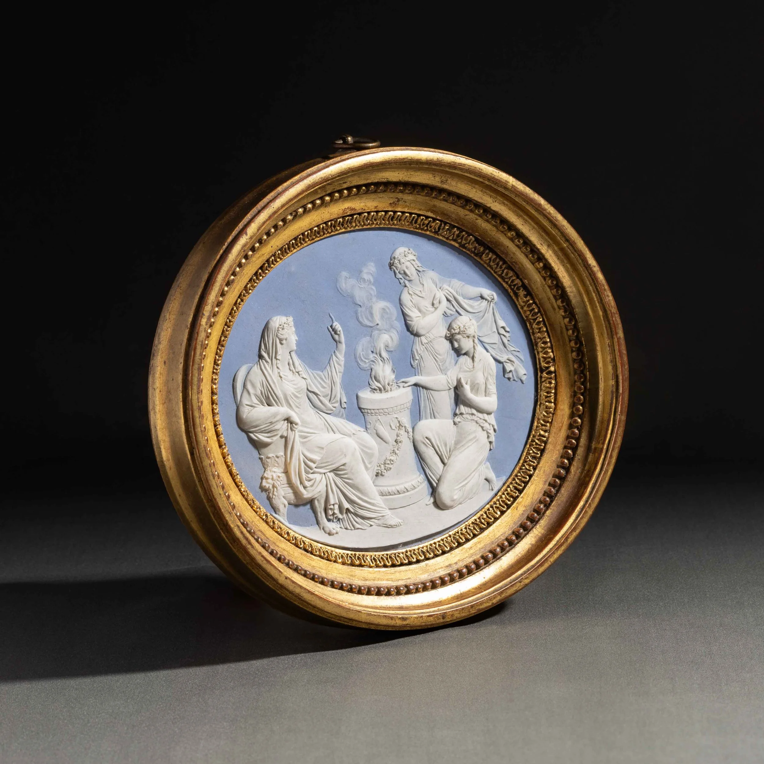 Une montre ancienne avec un cadran représentant une scène en relief en blanc, encadrée d'un cadre doré ornate, sur un fond noir.