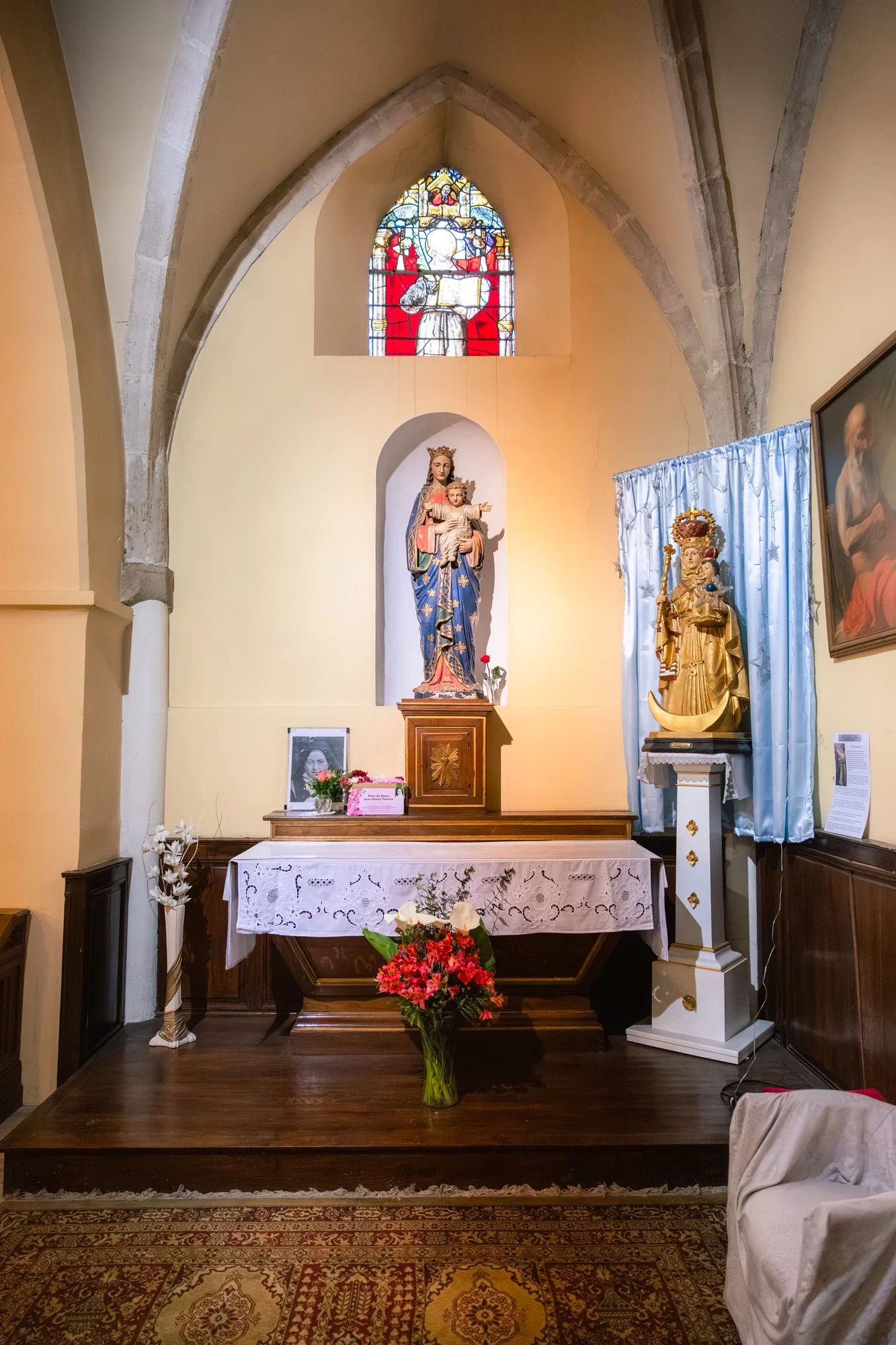 Une petite chapelle avec une statue de la Vierge Marie tenant l'enfant Jésus, décorée de fleurs, avec une peinture représentant un saint dans un cadre à droite, et un vitrail coloré en forme de croissant en haut.