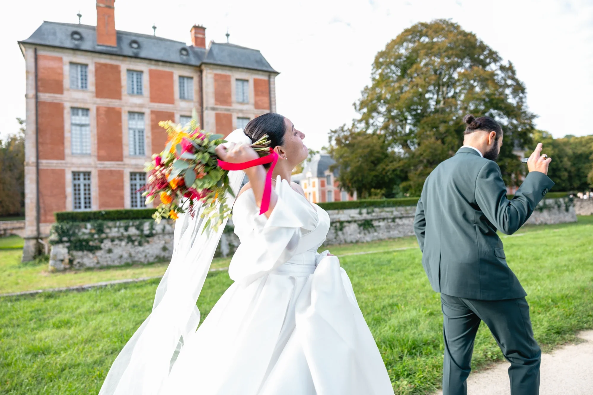 Une femme en robe de mariage avec un bouquet de fleurs colorées marche dans un jardin, accompagnée d'un homme en costume. Un grand arbre et un bâtiment ancien en arrière-plan.