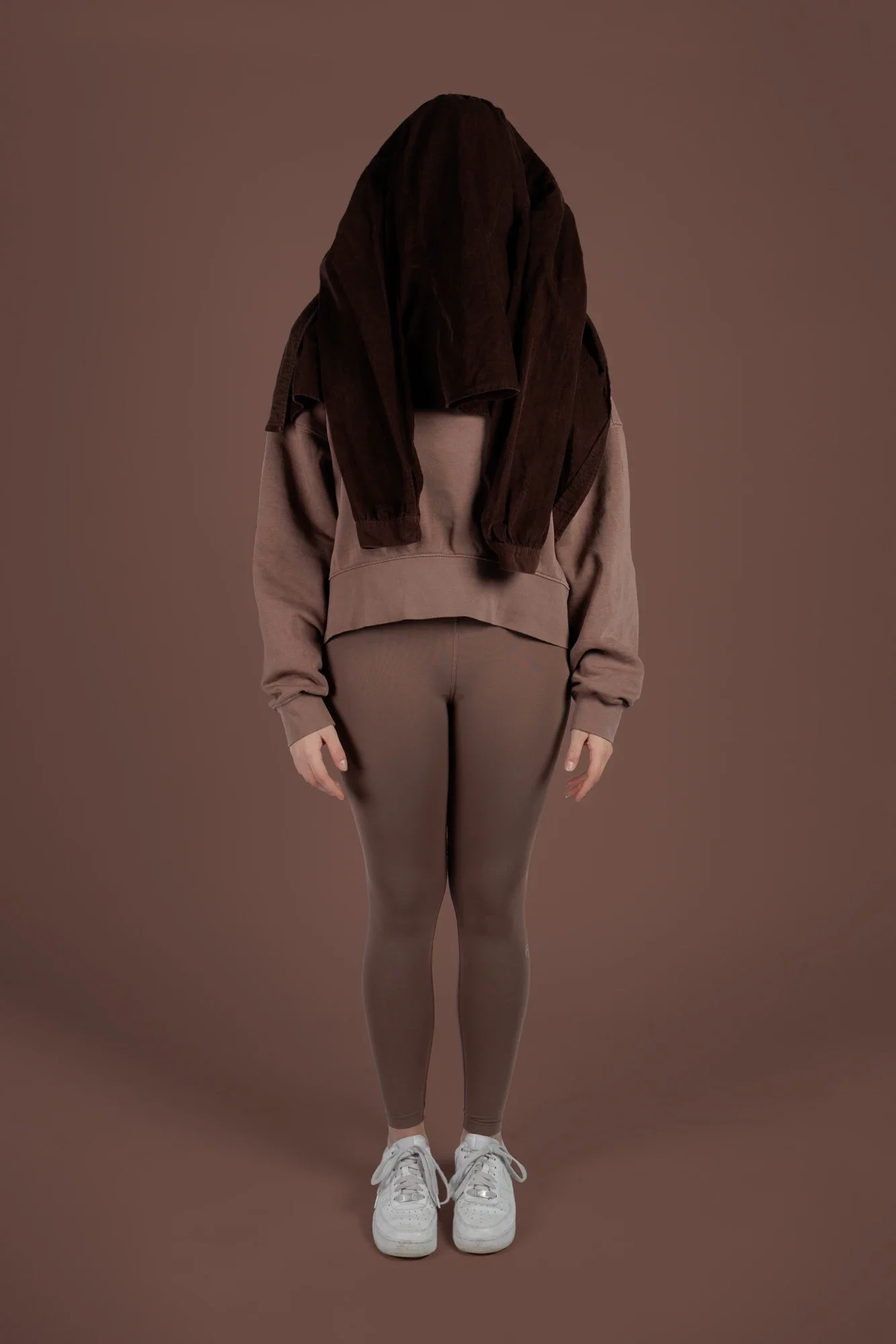 Une personne porte un sweat à capuche beige, un pantalon moulant beige et des baskets blanches, avec un sweat marron noué autour du cou et du dos, contre un fond marron.