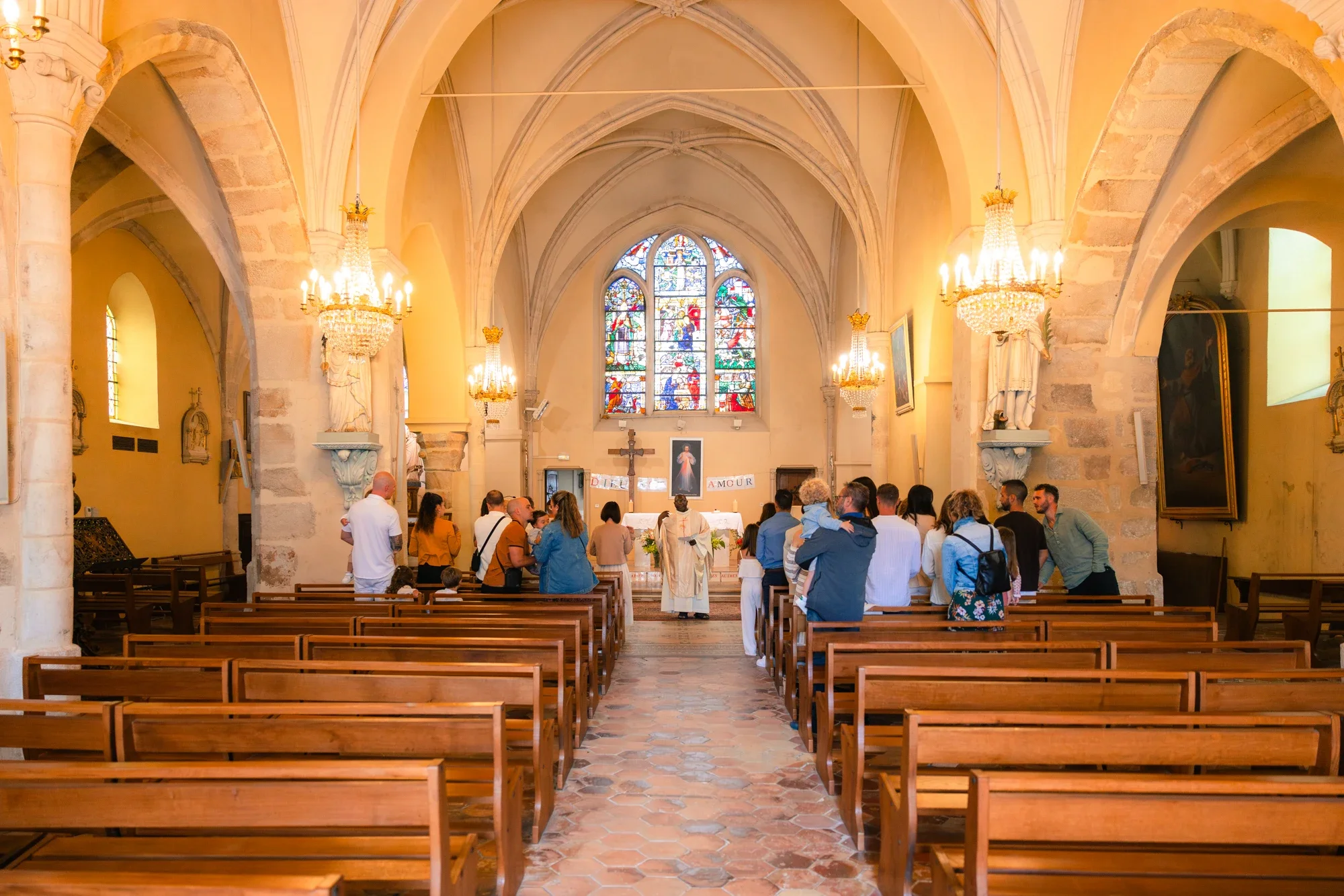 L'intérieur d'une église où des personnes assistent à une cérémonie religieuse. La scène est éclairée par des chandeliers et un vitrail coloré au fond.