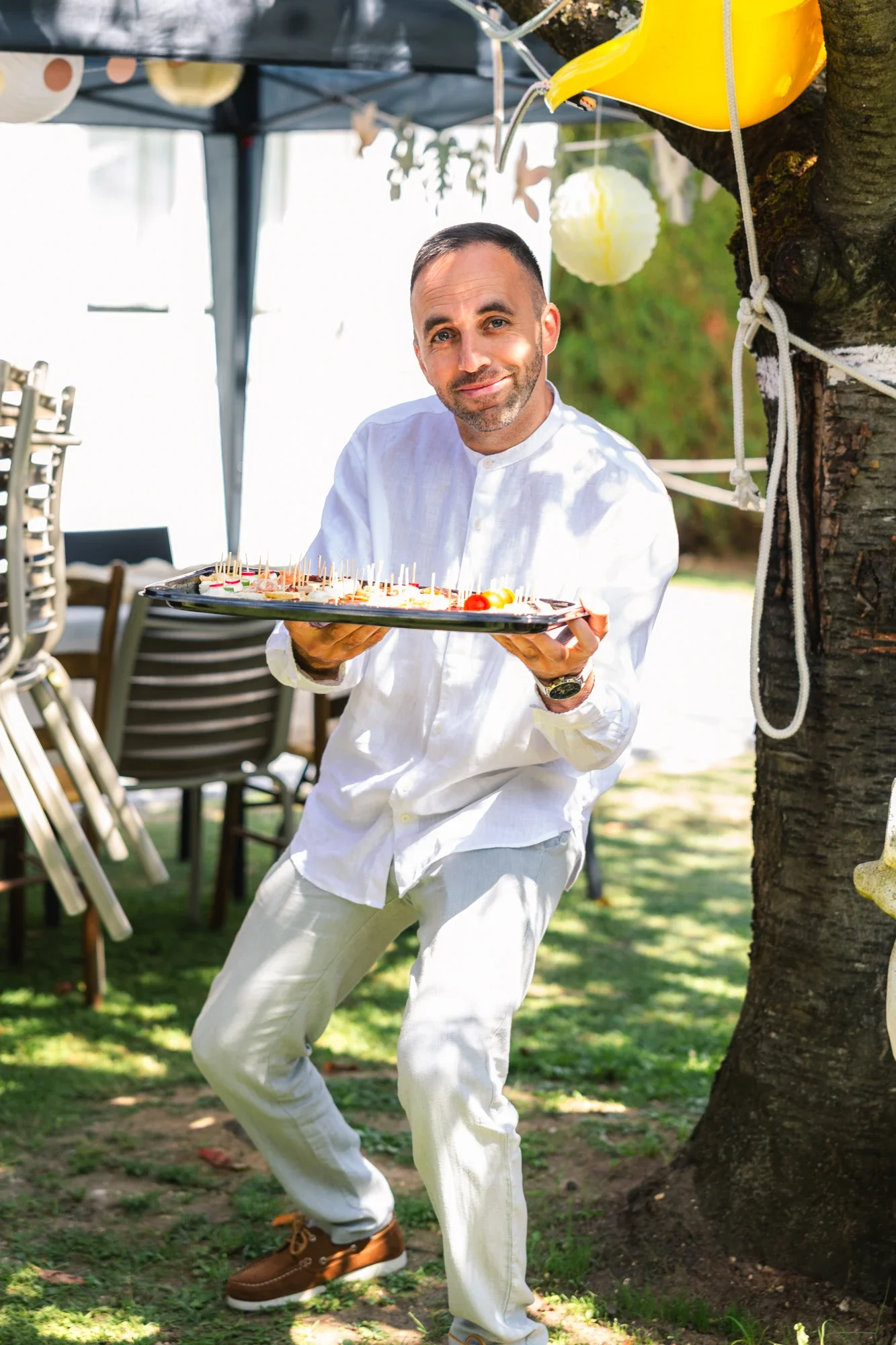 Un homme souriant porte un plateau avec des amuse-gueules lors d'une fête en plein air, attaché à un arbre avec des décorations colorées autour, sous un chapiteau.