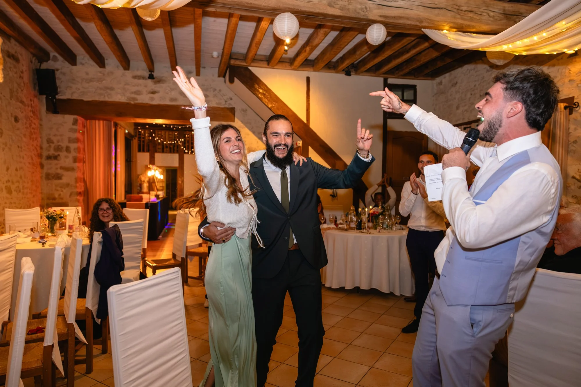 Groupe de personnes à une fête ou un mariage, joyeuses, dans une salle décorée d'éclairage chaud. Une femme et un homme souriants dansent et chantent avec un animateur ou un maître de cérémonie, tenant un microphone.