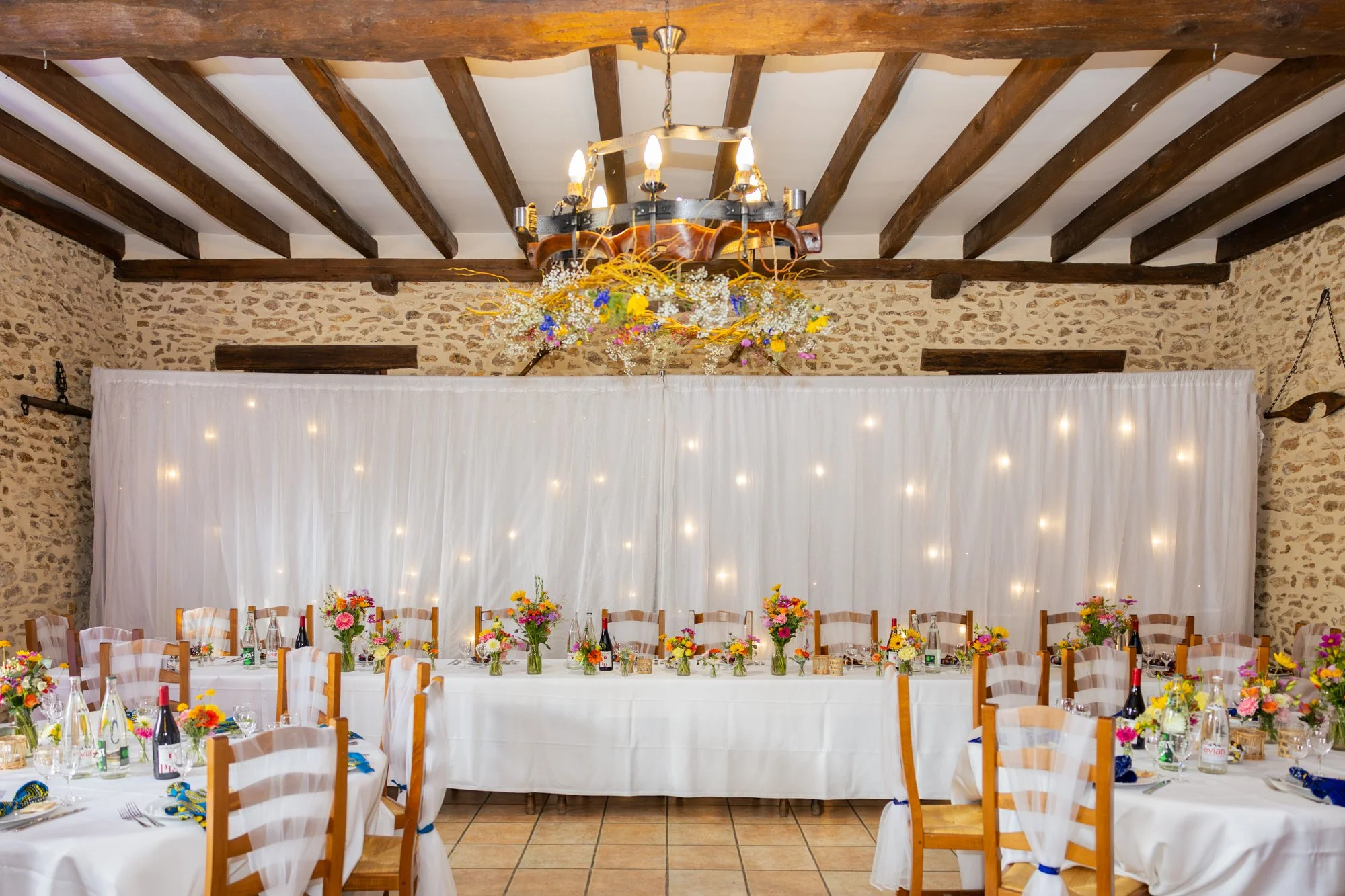 Salle de mariage décorée avec des tables rondes, des chaises en bois avec des housses blanches, des bouquets de fleurs colorés, une toile blanche avec des lumières et un chandelier en haut avec des fleurs suspendues, dans une pièce avec un plafond en