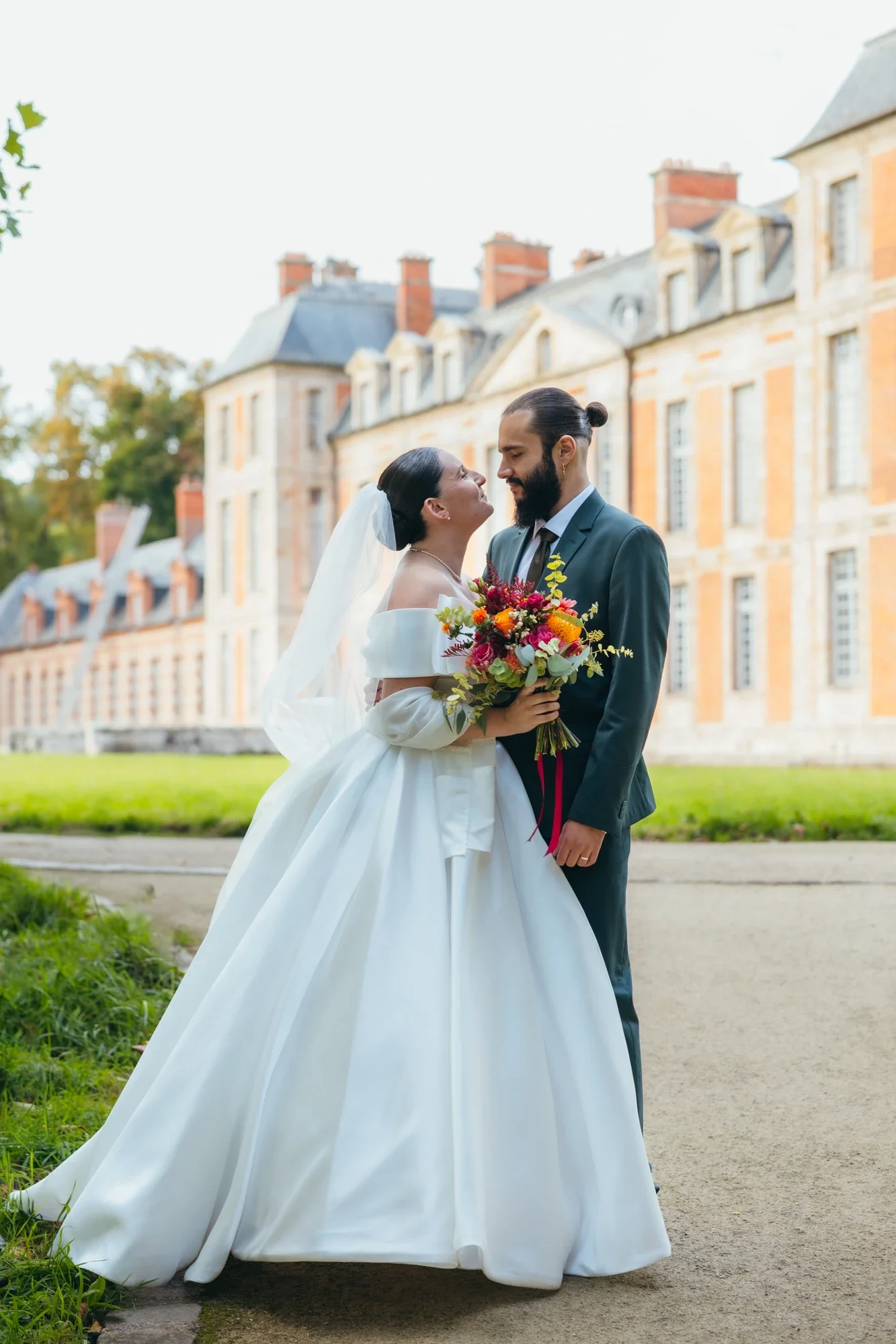 Un couple de mariés en costume et robe de mariage se regardant tendrement devant un bâtiment historique en pierre, avec le marié tenant un bouquet de fleurs colorées.