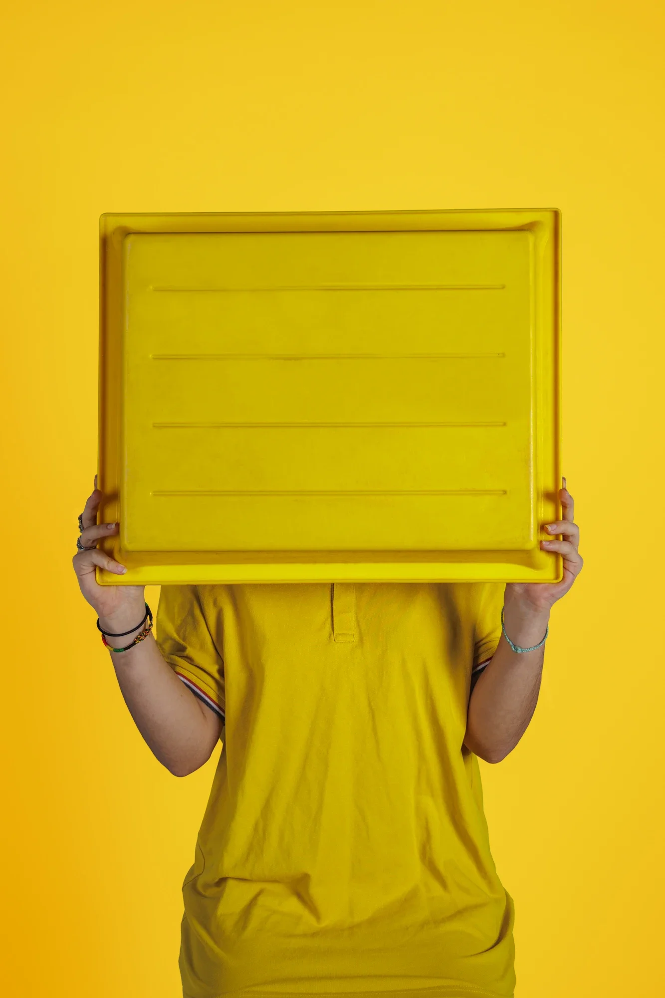 Personne en jaune tenant une grande boîte en plastique jaune devant le visage. Fond jaune.