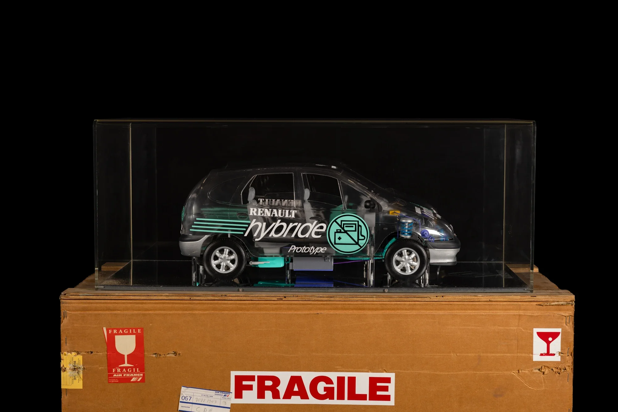 Modèle réduit de voiture électrique Renault hybride en prototype, exposé dans une boîte en plastique sur un socle en bois, avec des étiquettes de conformité et de sécurité.