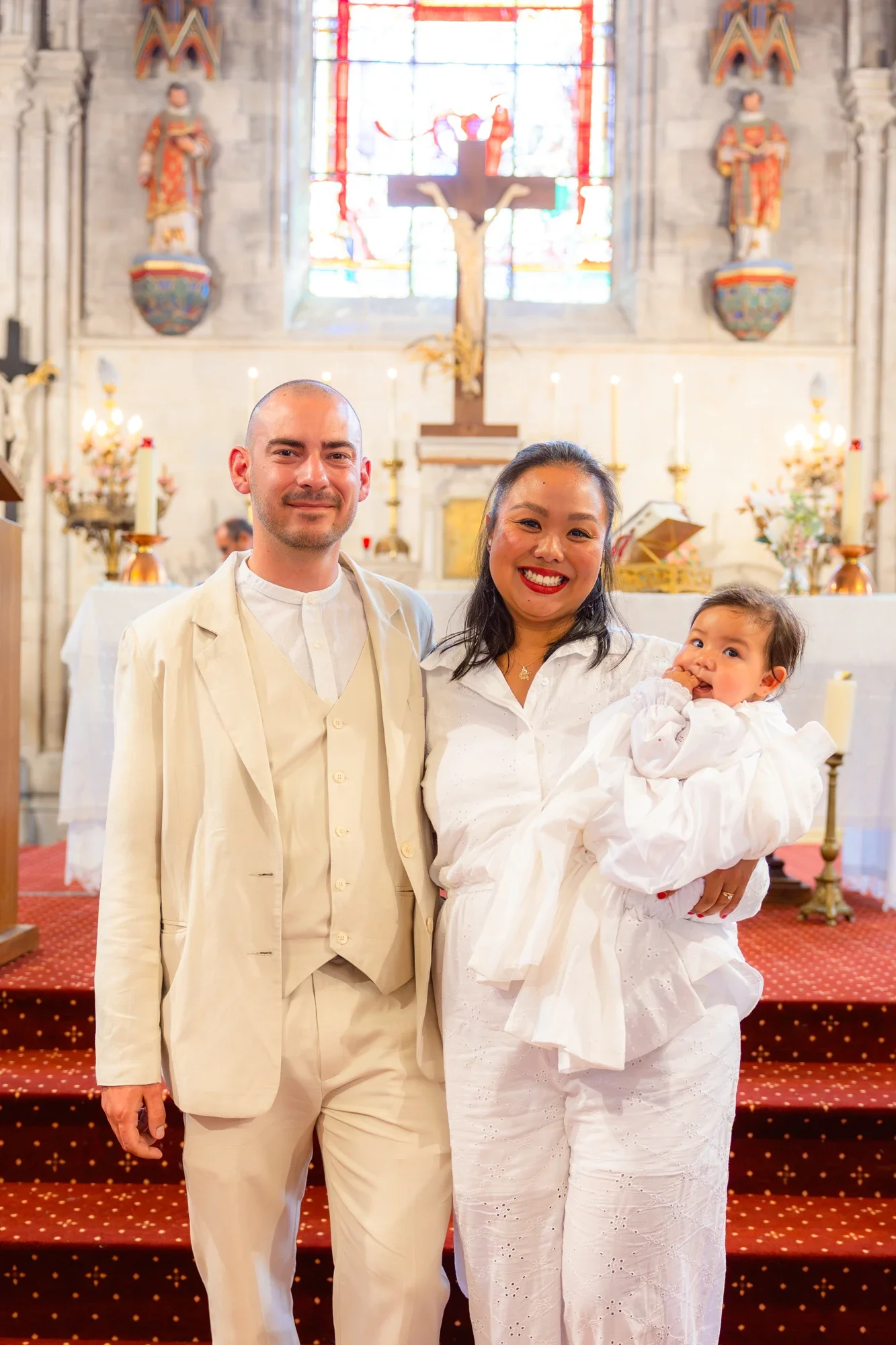 Photo d'une famille lors d'une cérémonie religieuse, avec un homme, une femme tenant un bébé, dans une église avec un autel et un crucifix en arrière-plan.