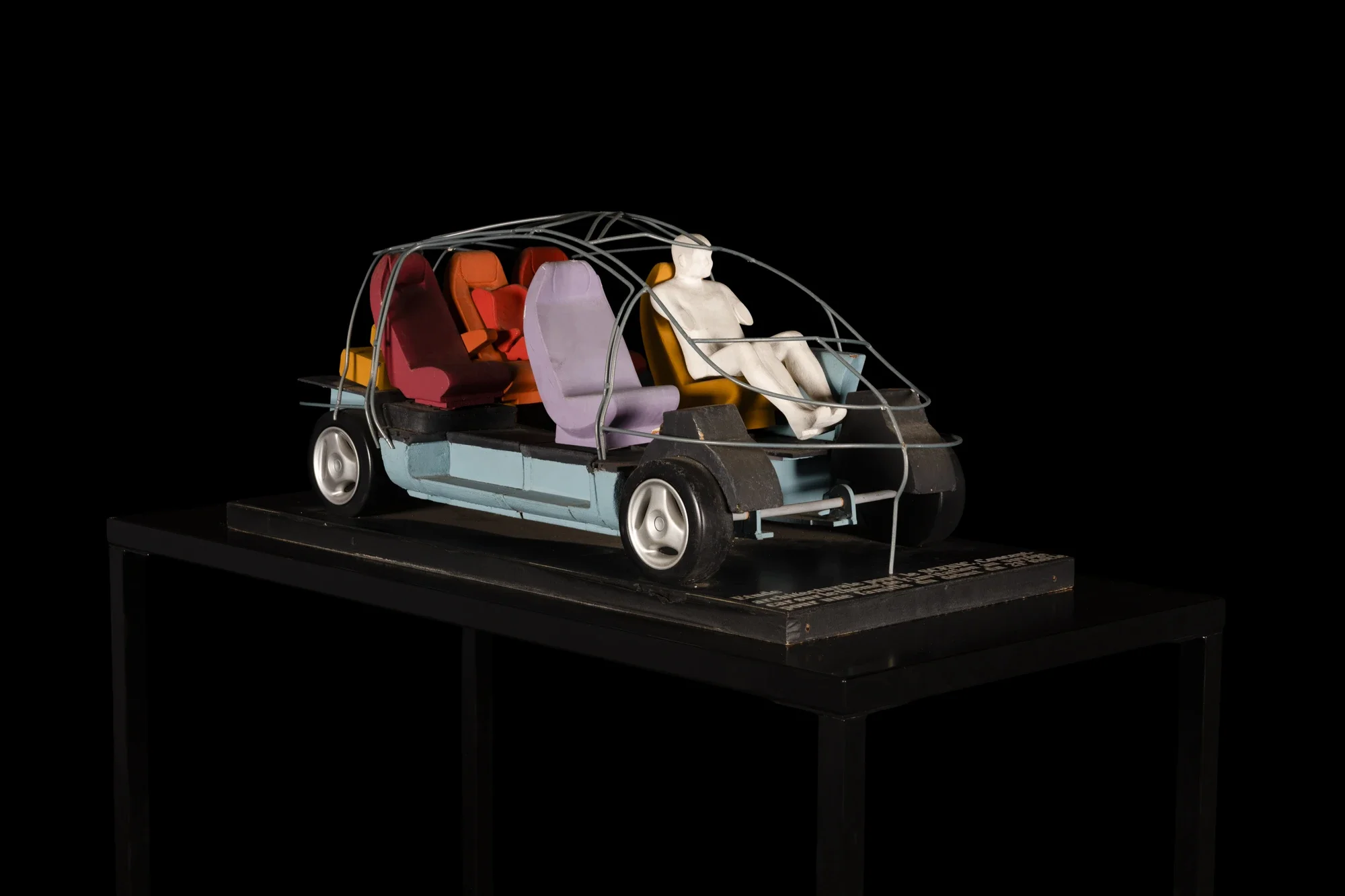 Miniature de voiture avec plusieurs sièges colorés et une figure humaine en plastique à l'avant, exposée sur un socle en noir.