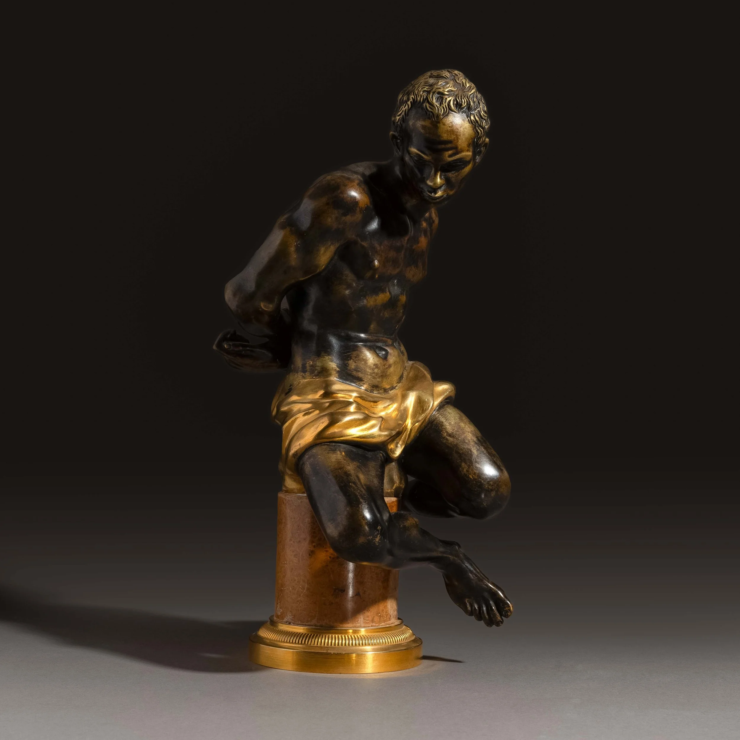 Sculpture en bronze d'un jeune enfant assis, regardant vers le bas, avec un décor en or au niveau du bassin. La sculpture est placée sur une base en bois.