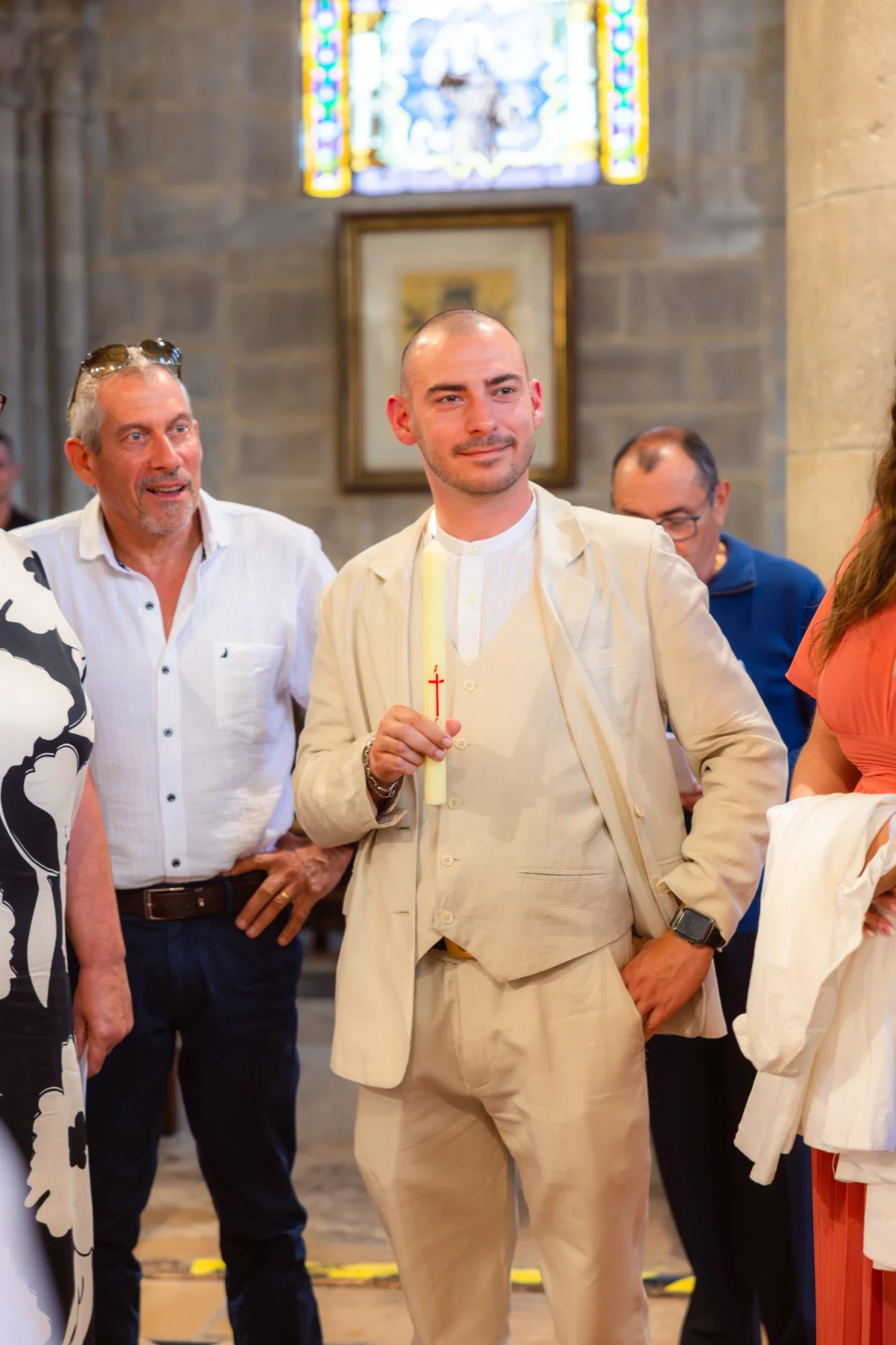 Un homme en costume clair tient une bougie dans une église, entouré de plusieurs personnes, avec un vitrail coloré en arrière-plan.