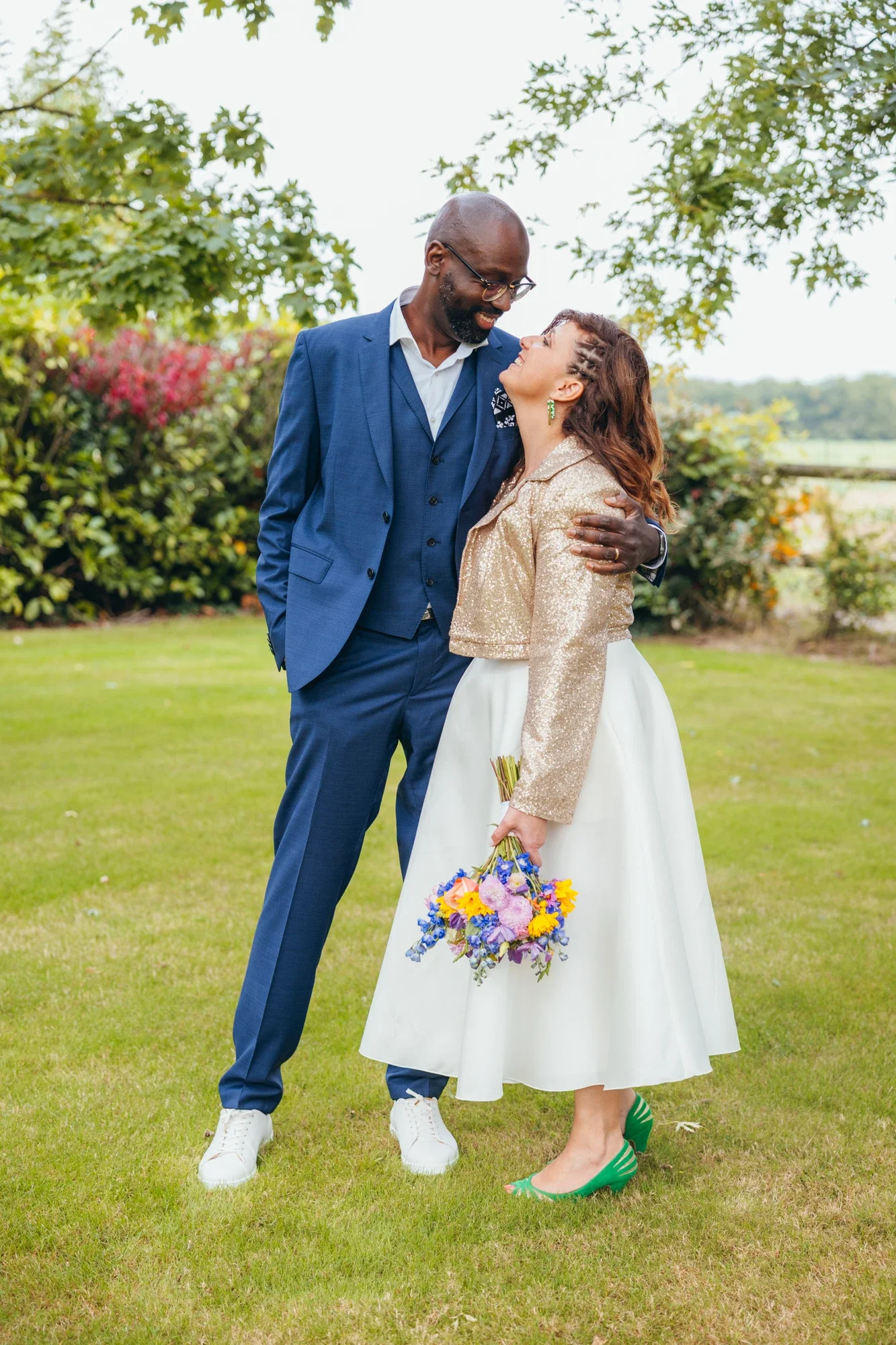 Un couple lors d'une célébration en plein air, lui en costume bleu, elle en robe blanche avec un haut beige scintillant, elle tient un bouquet de fleurs colorées, ils semblent heureux et s'embrassent tendrement.