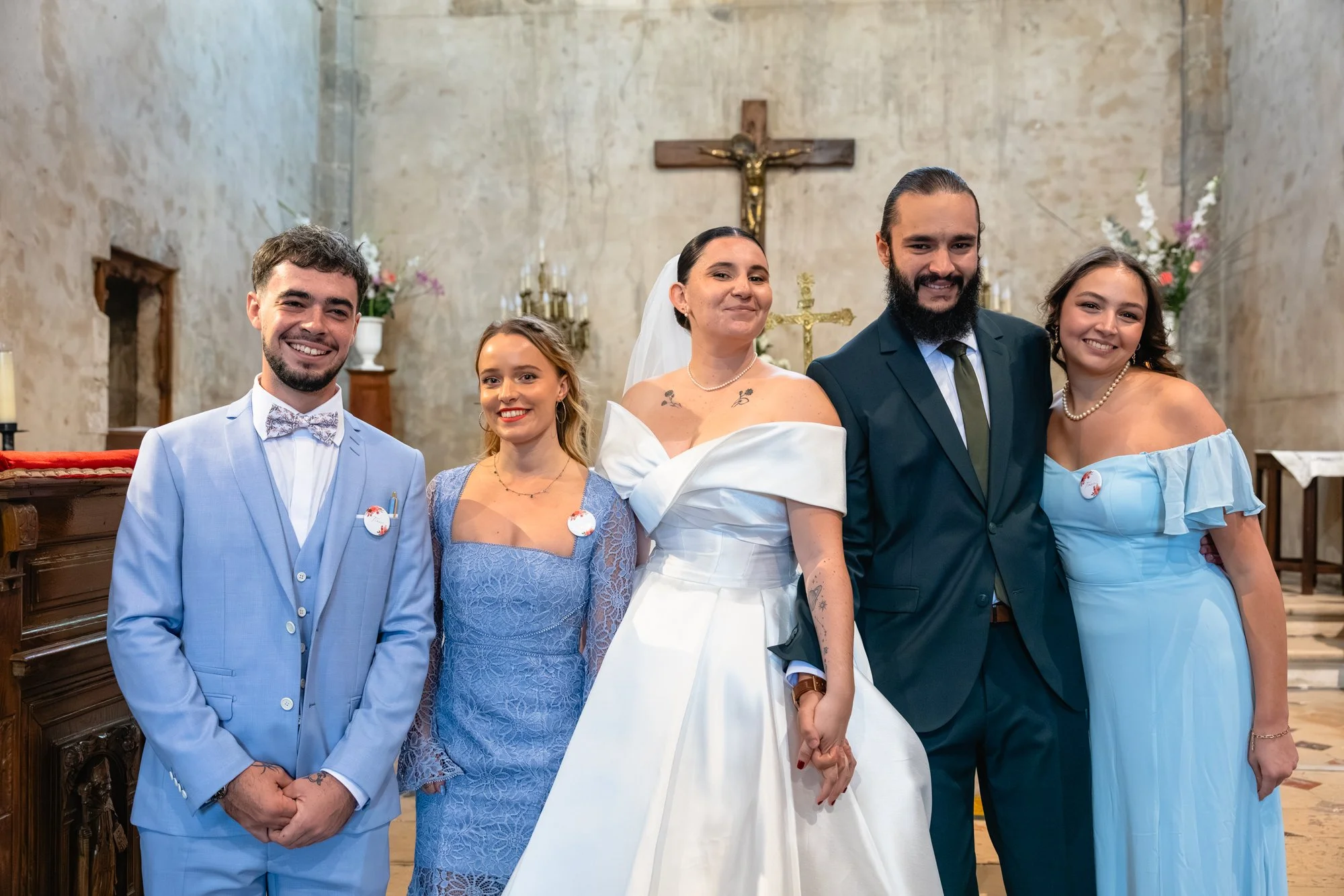 Groupe de cinq personnes lors d'une cérémonie religieuse, debout dans une église avec une croix en arrière-plan. La mariée en robe blanche au centre, entourée de deux femmes et deux hommes en costume ou robe élégante.