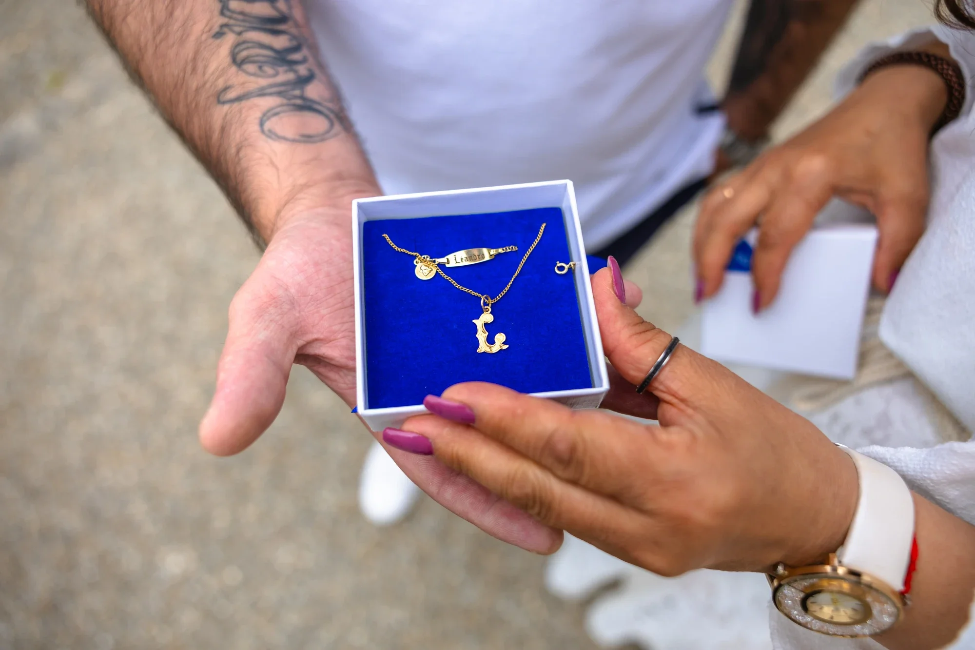 Une personne tient une boîte avec un collier en or et des pendentifs, dont un en forme de lettre 'L' et un autre en forme de lune, lors d'une occasion spéciale.
