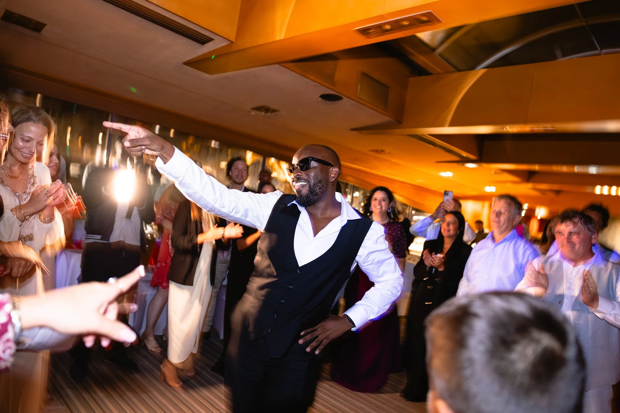 Un homme portant des lunettes de soleil et un costume sans manche danse au centre d'une fête elegant dans une salle avec un plafond en bois. Il est entouré de personnes qui l'applaudissent et sourient, la scène est lumineuse et festive.