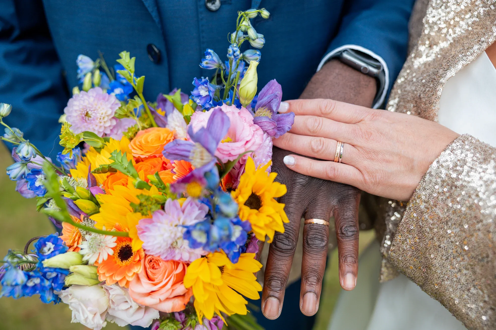 Deux personnes portent des alliances lors d'un mariage, une tenant un bouquet de fleurs colorées avec des roses, des tournesols et des fleurs violettes, et leur main est posée l'une sur l'autre.