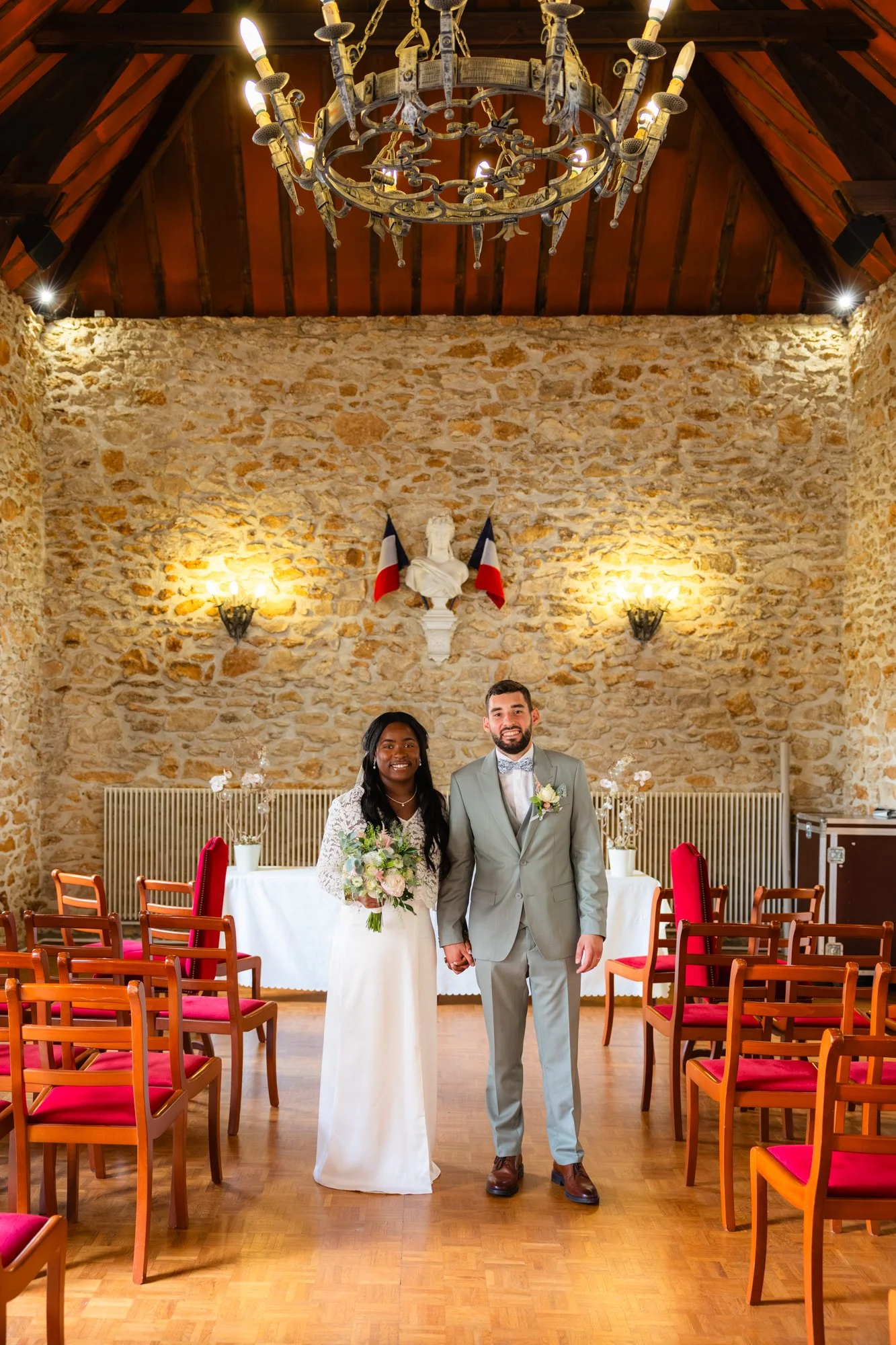 Photo d'un couple marié, debout dans une salle de réception avec un mur en pierre, des chaises en bois avec assises rouges et une table décorée. La bride porte une robe blanche, le marié un costume gris clair. Il y a des drapeaux français derrière eu