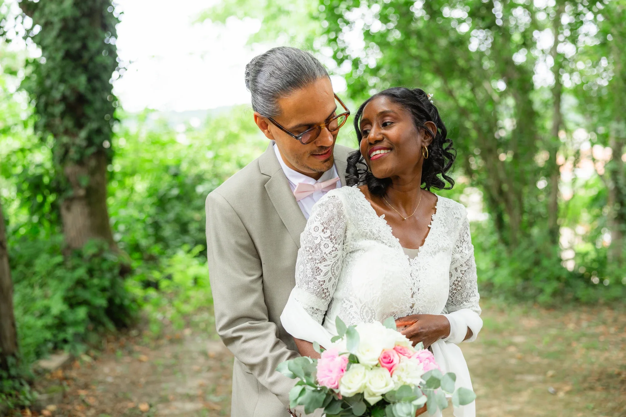 Un couple de mariés dans un parc, la femme tenant un bouquet de fleurs roses et blanches, l'homme en costume clair, tous deux souriants, entourés d'arbres verts.