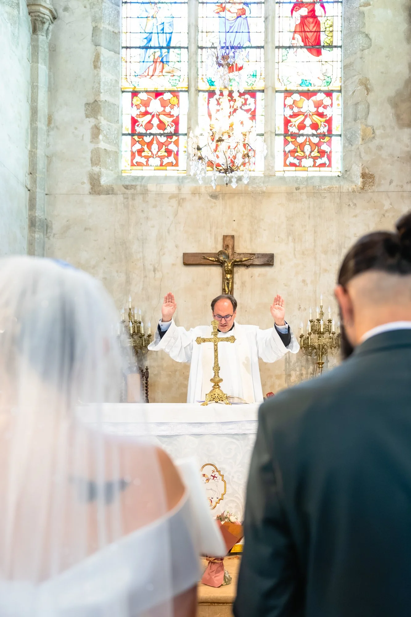 Une scène religieuse dans une église avec un prêtre célébrant une messe. Le prêtre porte des vêtements blancs et se tient devant un autel avec une croix fleurie. Au fond, un crucifix en bois avec la statue de Jésus crucifié, et une grande fenêtre en 