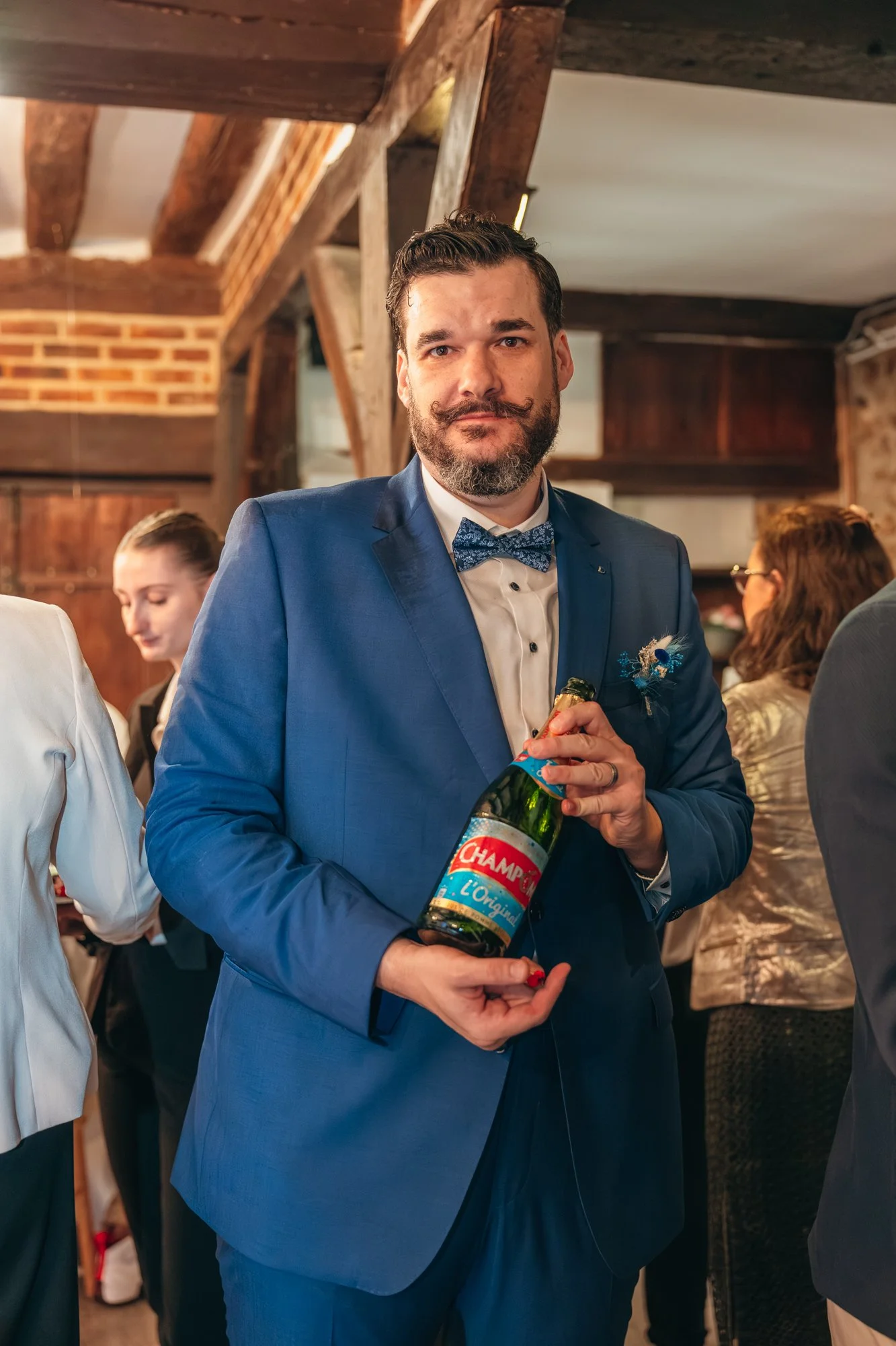 Un homme en costume bleu avec une cravate à motif tient une bouteille de champagne lors d'une célébration. Il a une barbe et une moustache, et l'ambiance indique une fête ou un événement social.