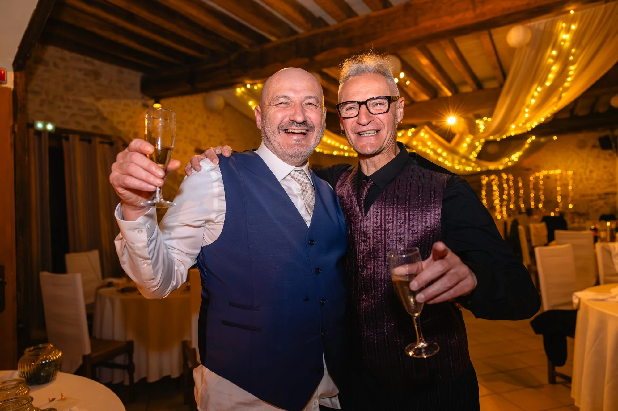 Deux hommes souriants tiennent des verres de champagne lors d'une fête dans une salle décorée avec des guirlandes lumineuses, ambiance chaleureuse et festive.