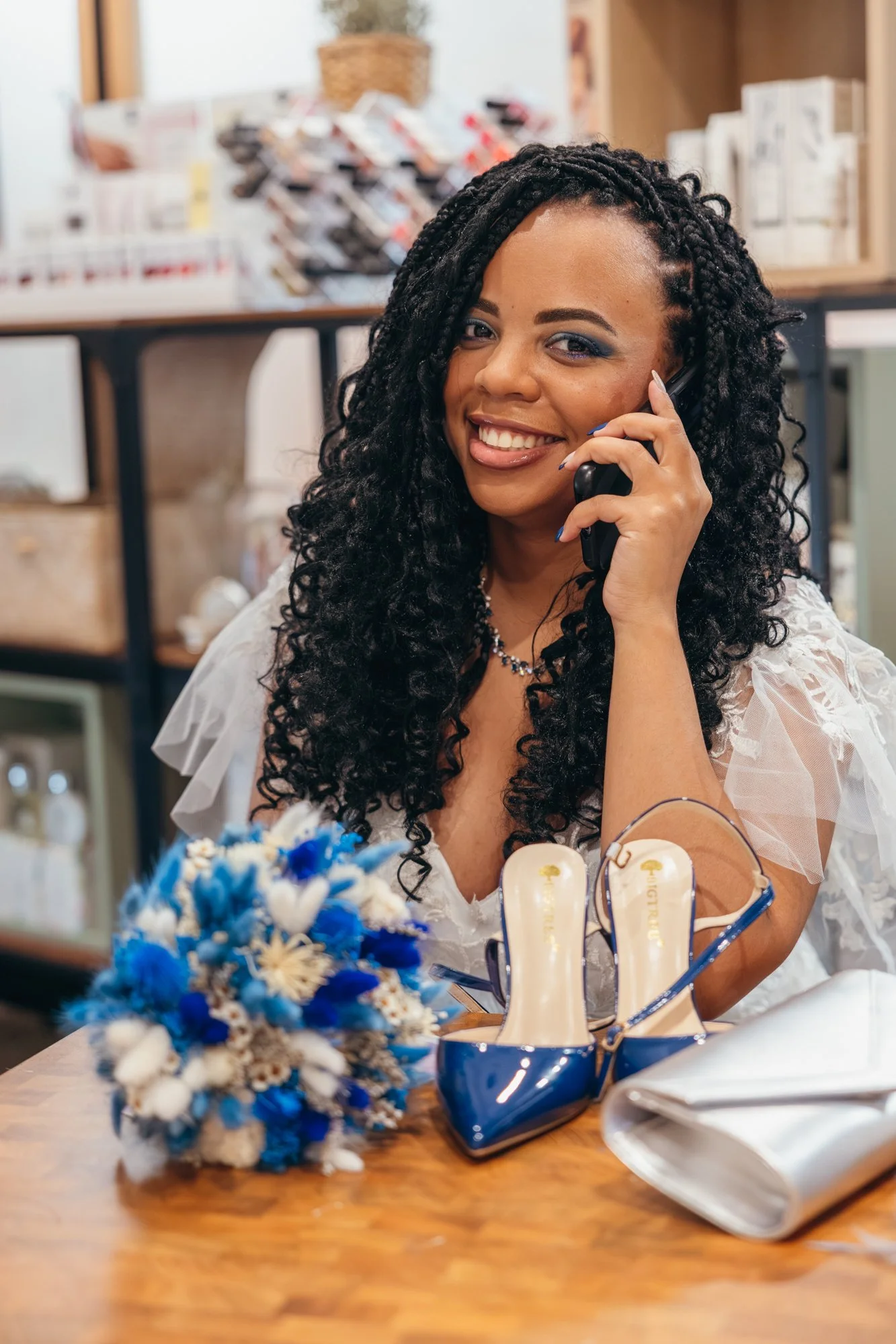 Une femme souriante avec une coiffure tressée noire, portant un maquillage bleu, parle au téléphone dans un magasin. Des chaussures bleues et une bouquet de fleurs bleues sont sur la table.