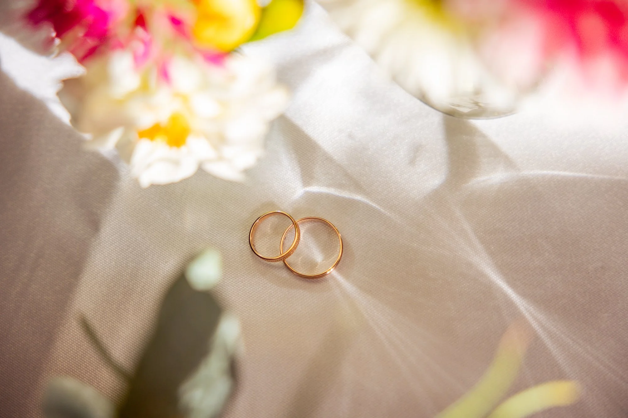 Deux alliances en or posées sur une surface en satin, entourées de fleurs blanches et roses avec de la lumière douce et un fond flou.