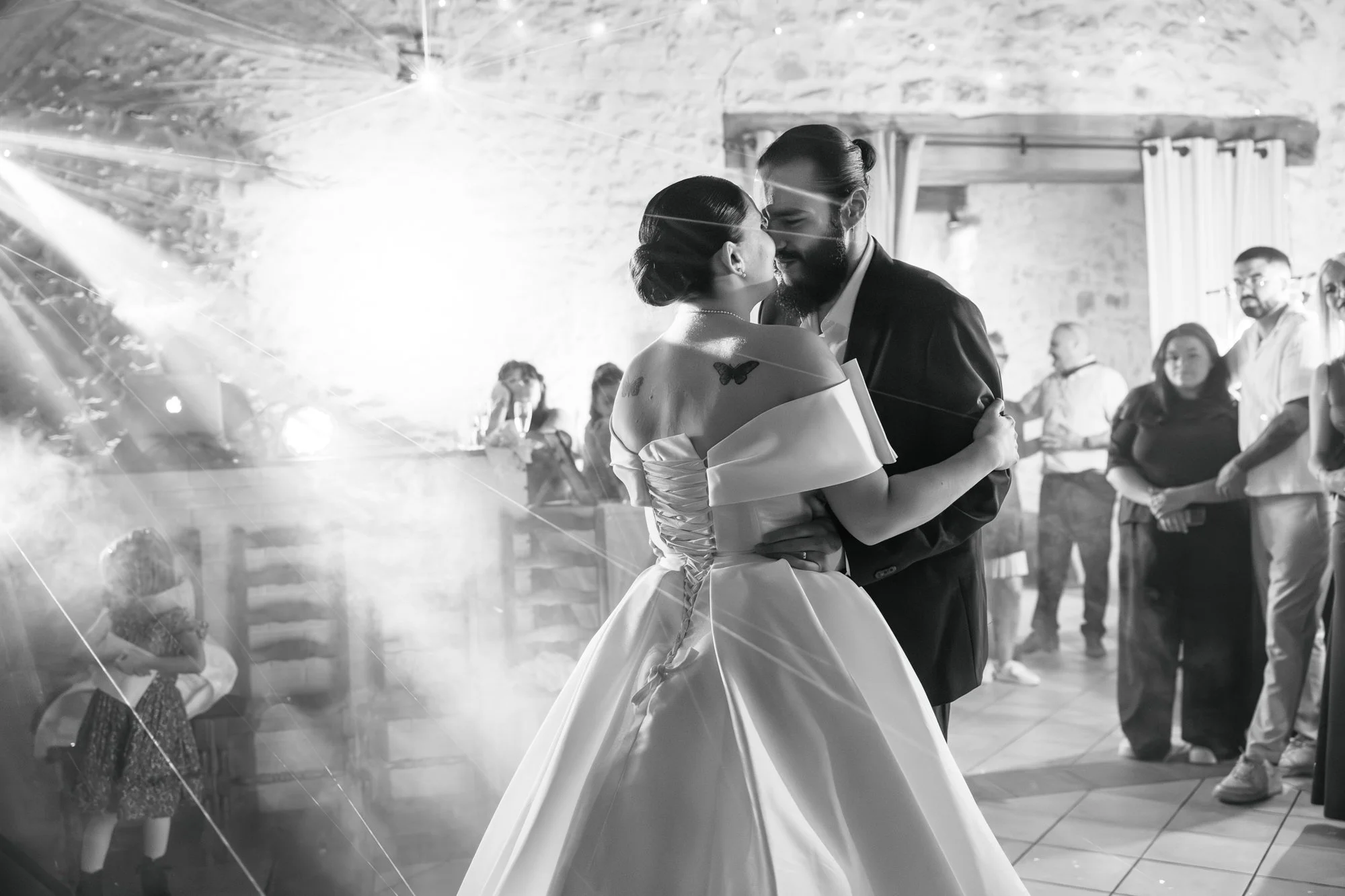 Un couple danse lors d'une célébration, entouré d'invités dans une pièce avec murs en pierre et rideaux blancs.